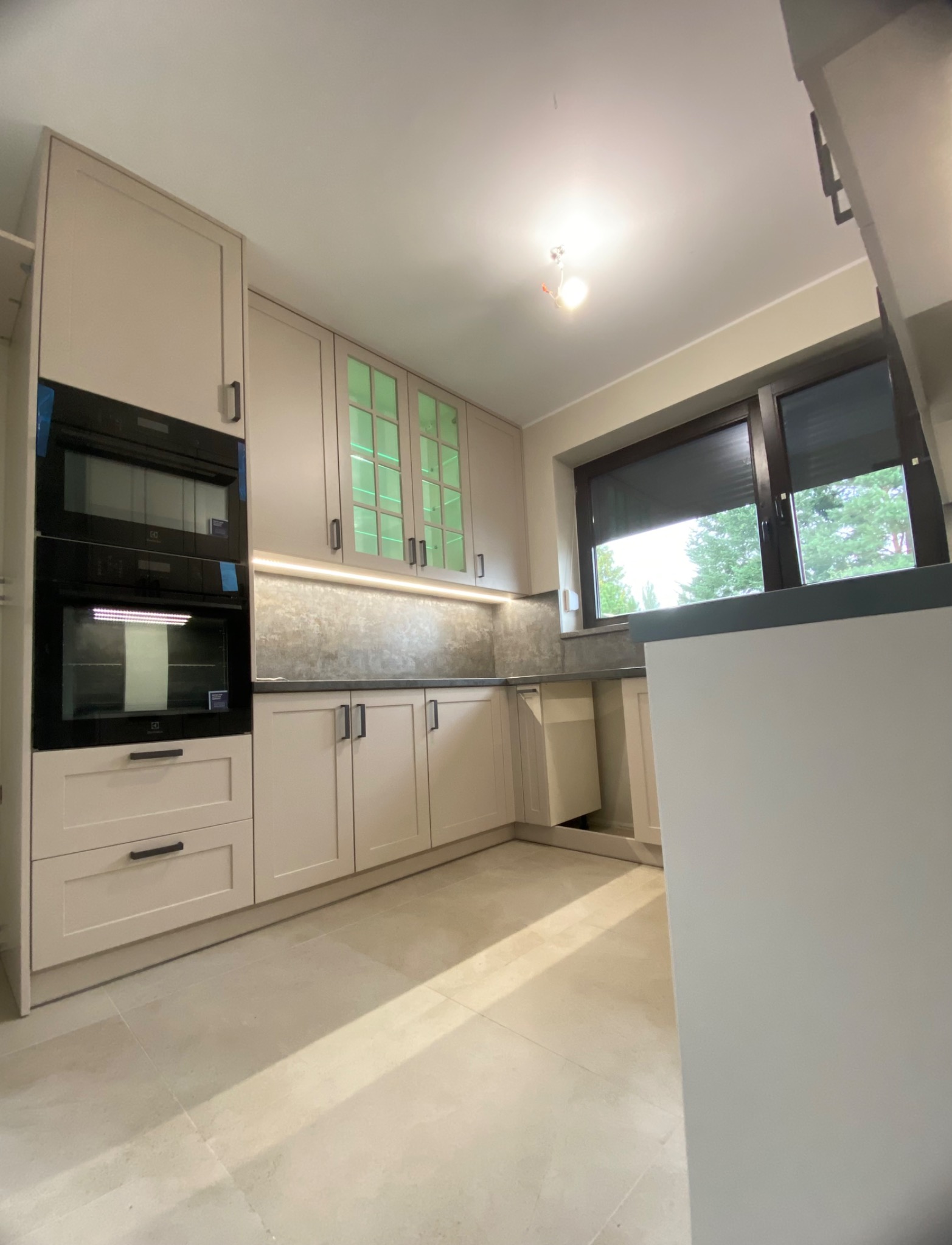 Jasna kuchnia z zabudową, z piekarnikiem w zabudowie i szafkami z zielonymi szybami. Nad blatem oświetlenie LED. Widok na okno z roletami.