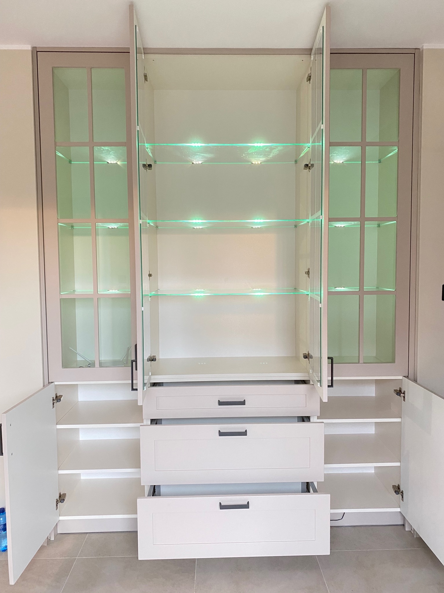 Biała szafa z szklanymi drzwiczkami i podświetlanymi półkami, otwarte drzwiczki i wysunięte szuflady, widoczne wnętrze szafy.