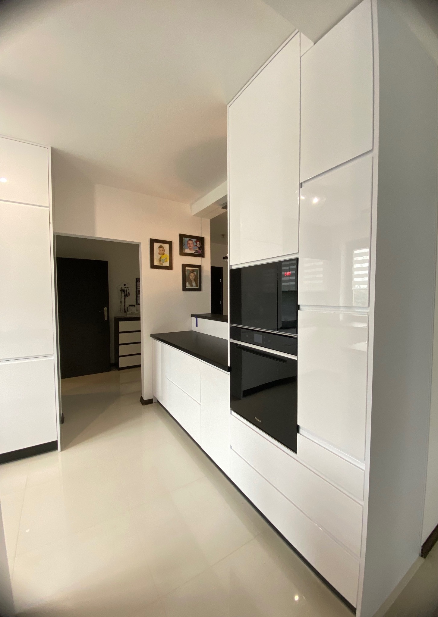 Biała zabudowa kuchenna z piekarnikiem i mikrofalówką w nowoczesnym wnętrzu. Kontrastowy czarny blat i sprzęt AGD. Widoczne zdjęcia rodzinne w tle.