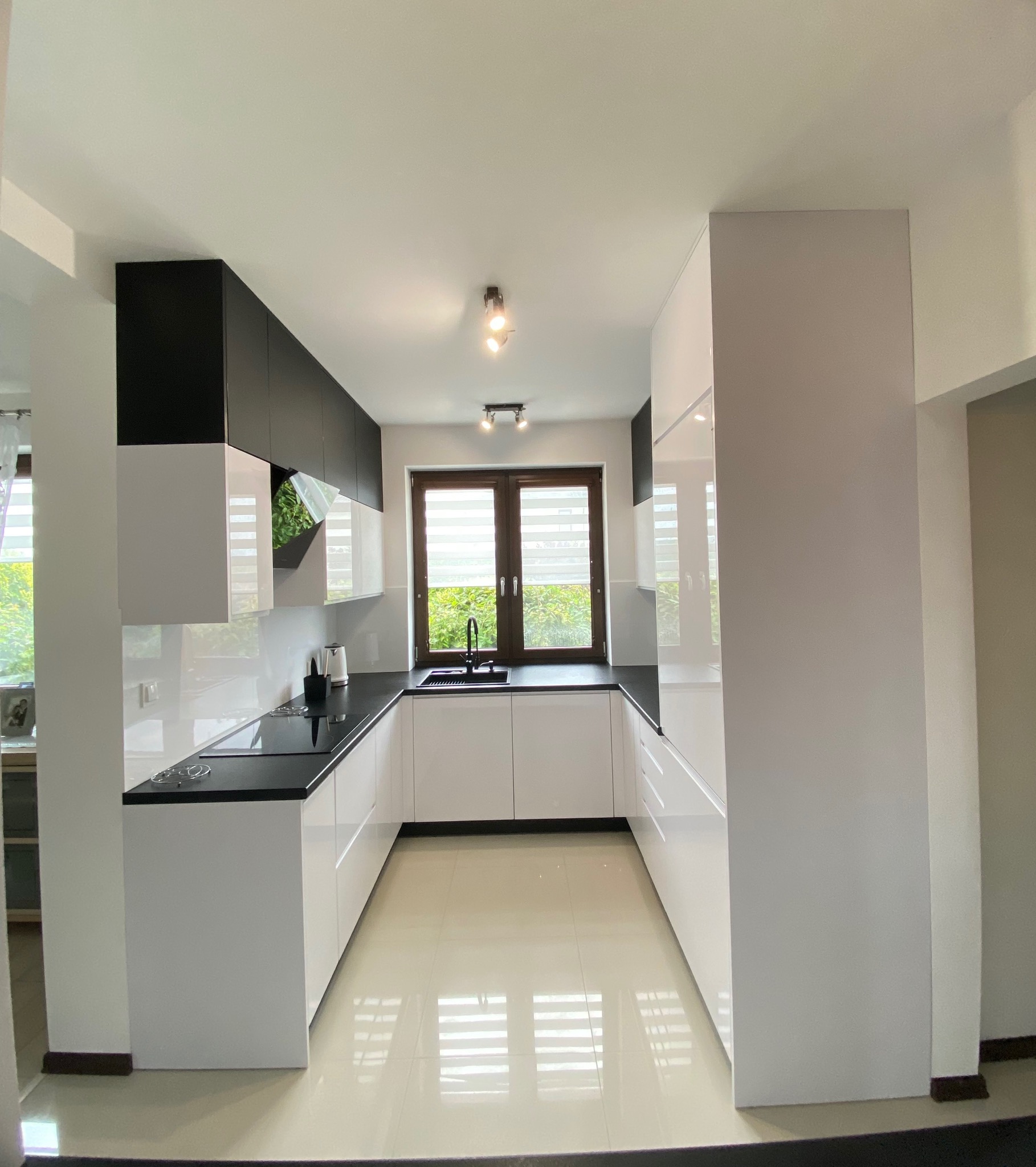 Nowoczesna kuchnia na wymiar z białymi frontami i czarnym blatem, z oknem w tle. Ułożona w kształcie litery U, z widoczną armaturą i elementami dekoracyjnymi.