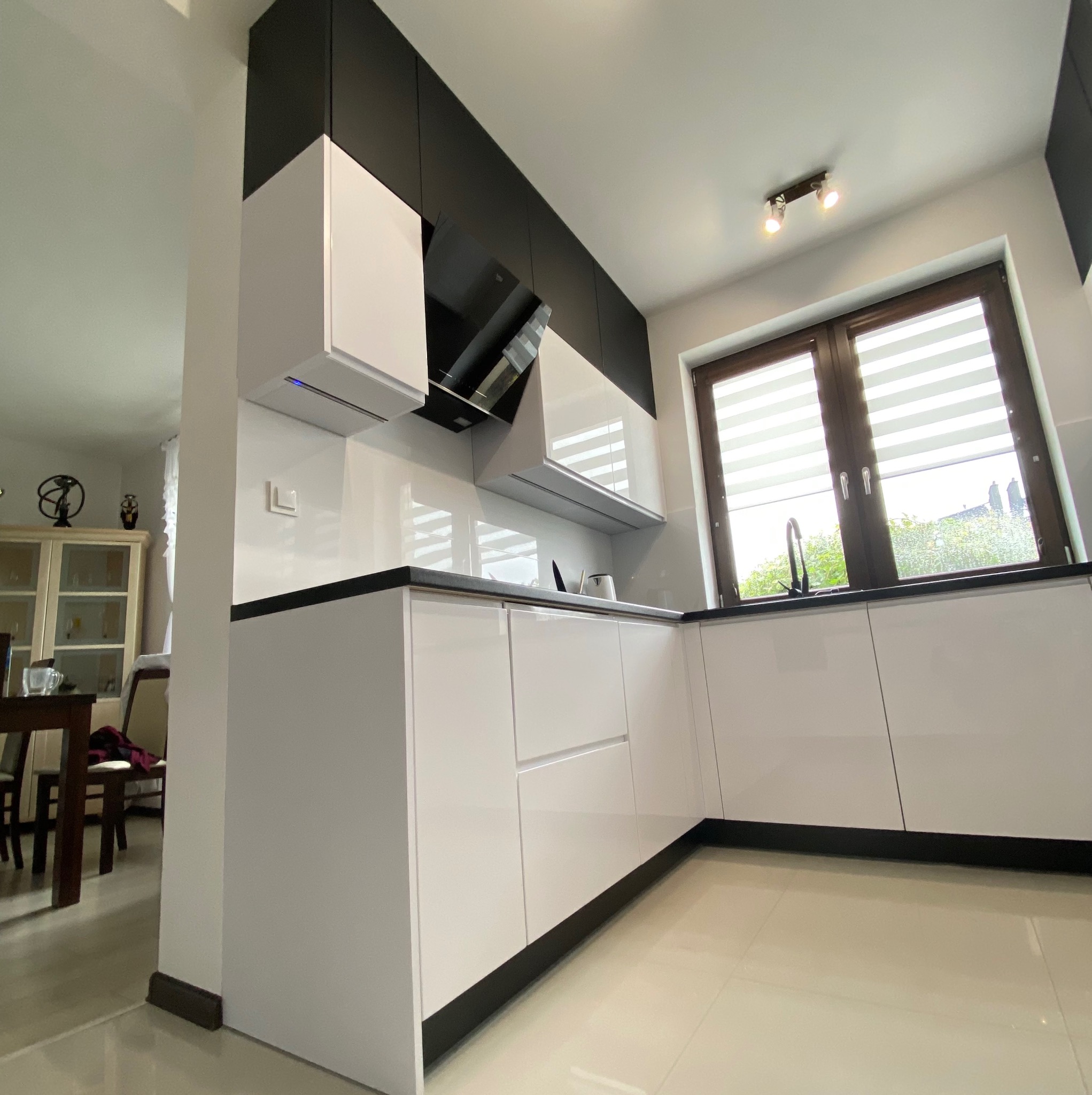 Biała kuchnia z czarnymi akcentami, błyszczące fronty, widok z dołu, okno z żaluzjami w tle, nowoczesny design.