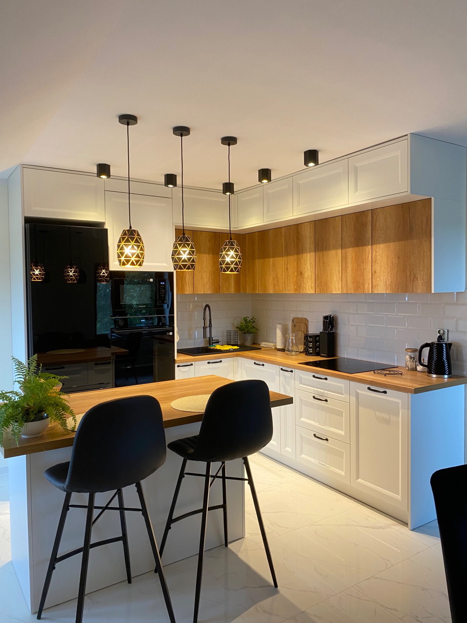 Nowoczesna kuchnia: białe szafki z drewnianymi akcentami, czarne sprzęty AGD i wyspa z hokerami na tle marmurowej podłogi, oświetlona wiszącymi lampami.