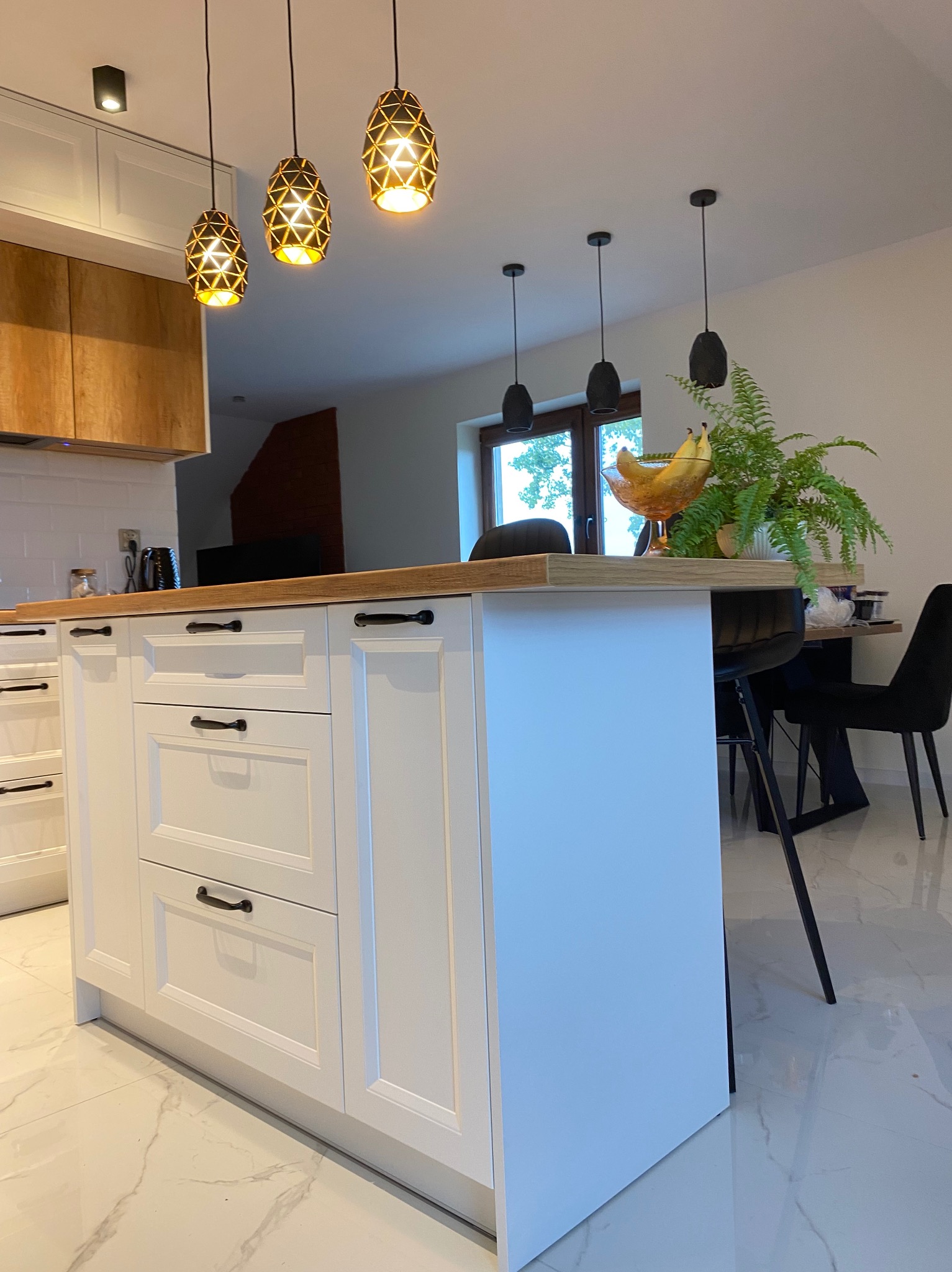 Biała wyspa kuchenna z drewnianym blatem i czarnymi uchwytami w nowoczesnej kuchni. Nad wyspą wiszą trzy złote lampy o geometrycznym wzorze.