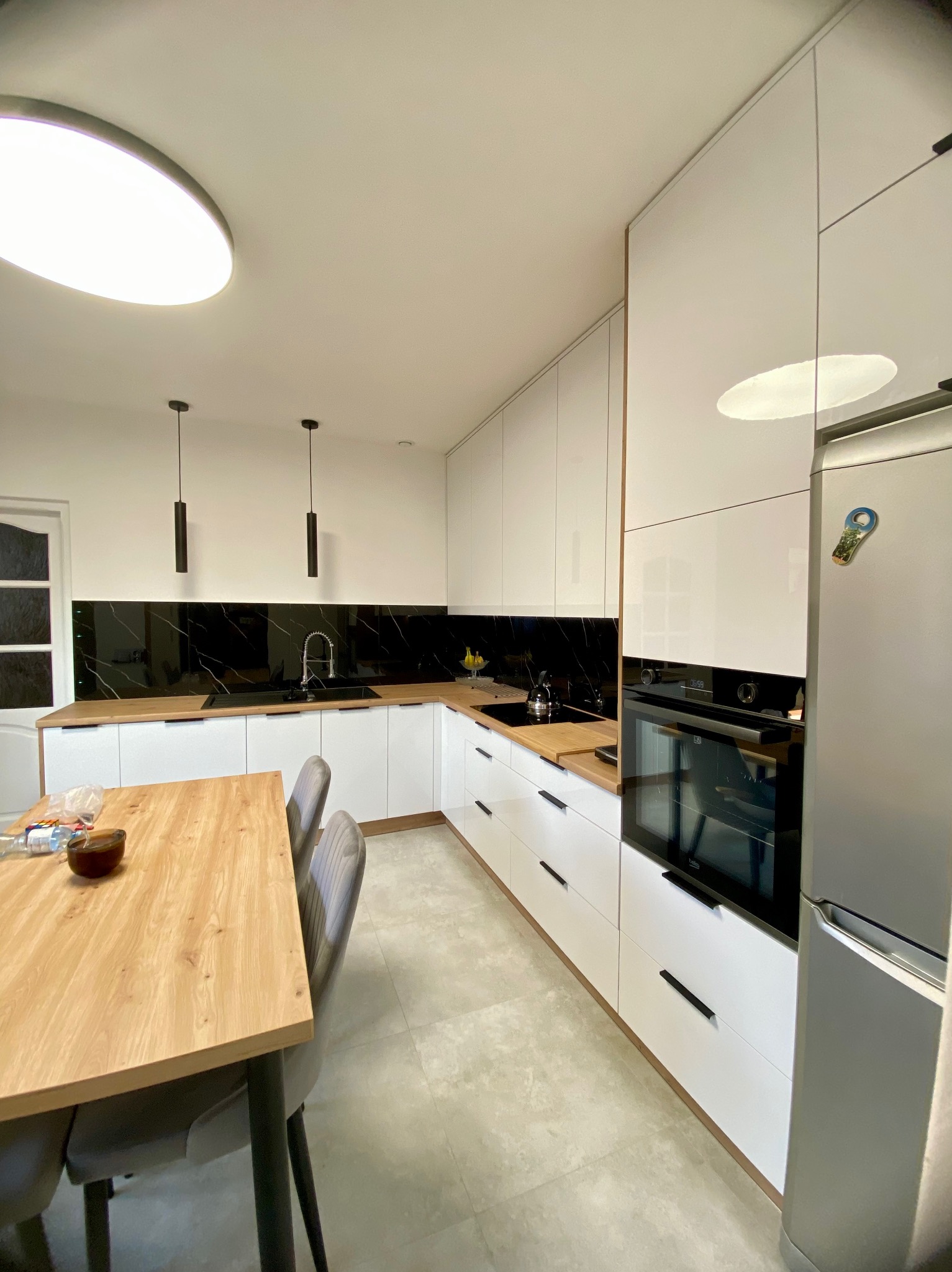 Nowoczesna kuchnia w bieli z czarnym blatem i drewnianym stołem. Minimalistyczne lampy wiszące i srebrna lodówka. Całość utrzymana w jasnej tonacji.