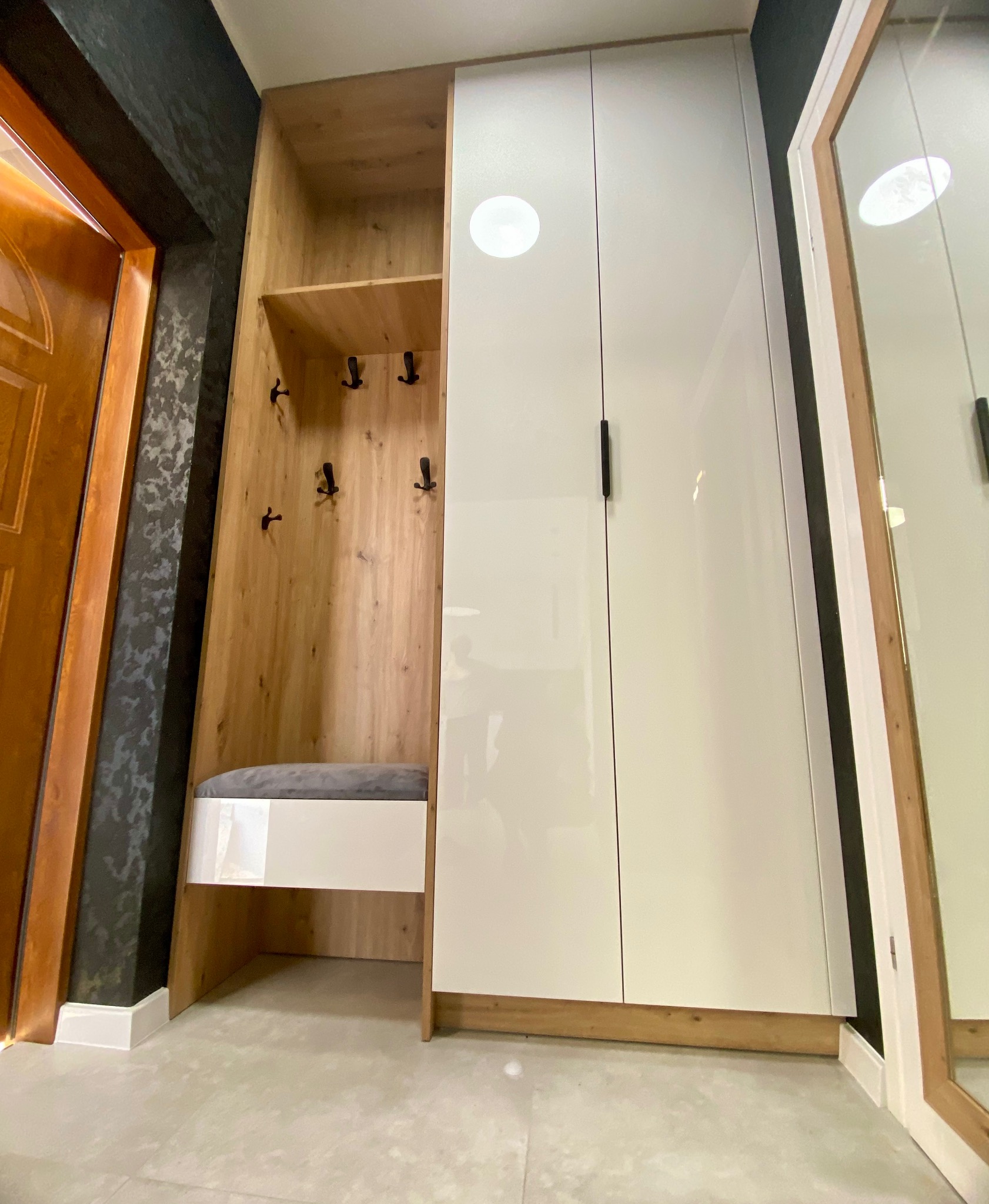 Funkcjonalna szafa w przedpokoju z siedziskiem i wieszakami, wykończona w drewnie i bieli, z lustrem po prawej stronie, na tle ciemnej ściany.