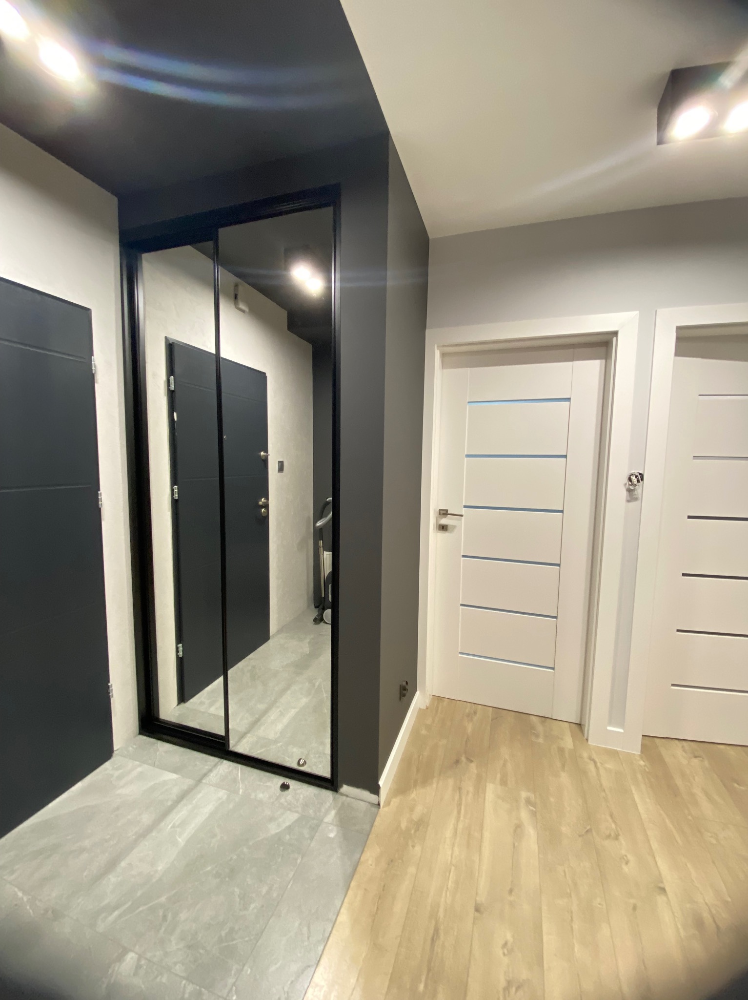 Szafa wnękowa z przesuwnymi lustrzanymi drzwiami w ciemnej ramie, widoczna w korytarzu z szarą podłogą i białymi drzwiami.