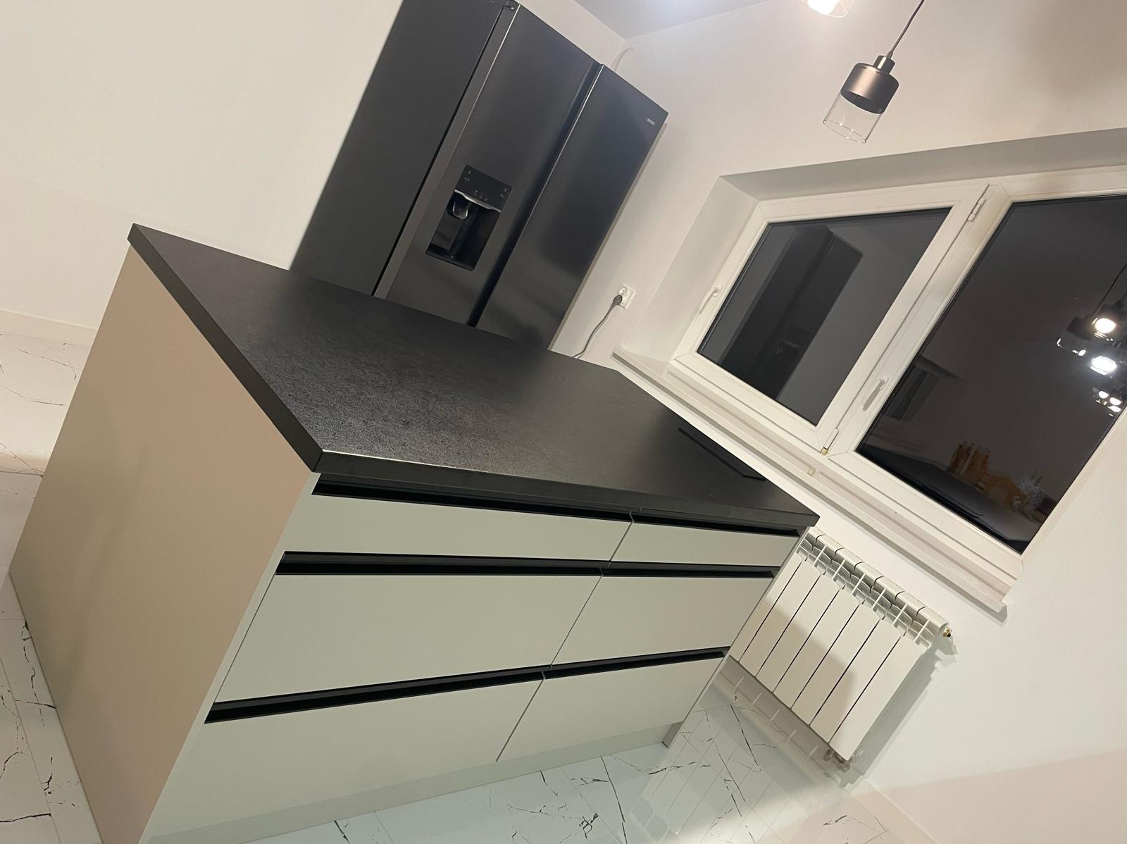 Nowoczesna wyspa kuchenna z szarym blatem, frontami w odcieniach beżu i czarnymi akcentami, na tle białej ściany z oknem i grzejnikiem.