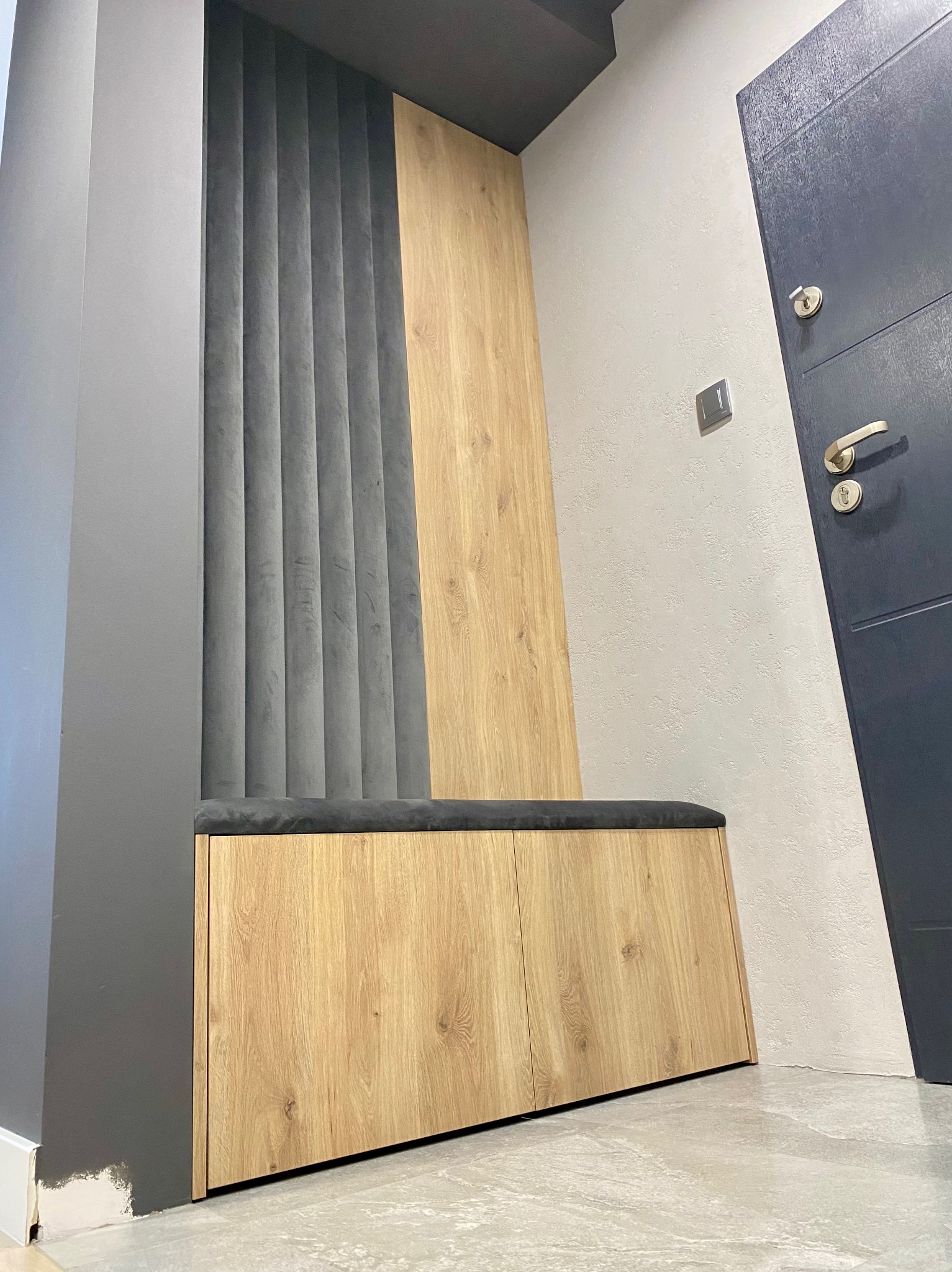 Nowoczesna szafka z siedziskiem i panelem ściennym w odcieniach szarości i drewna, umieszczona przy wejściu do mieszkania.