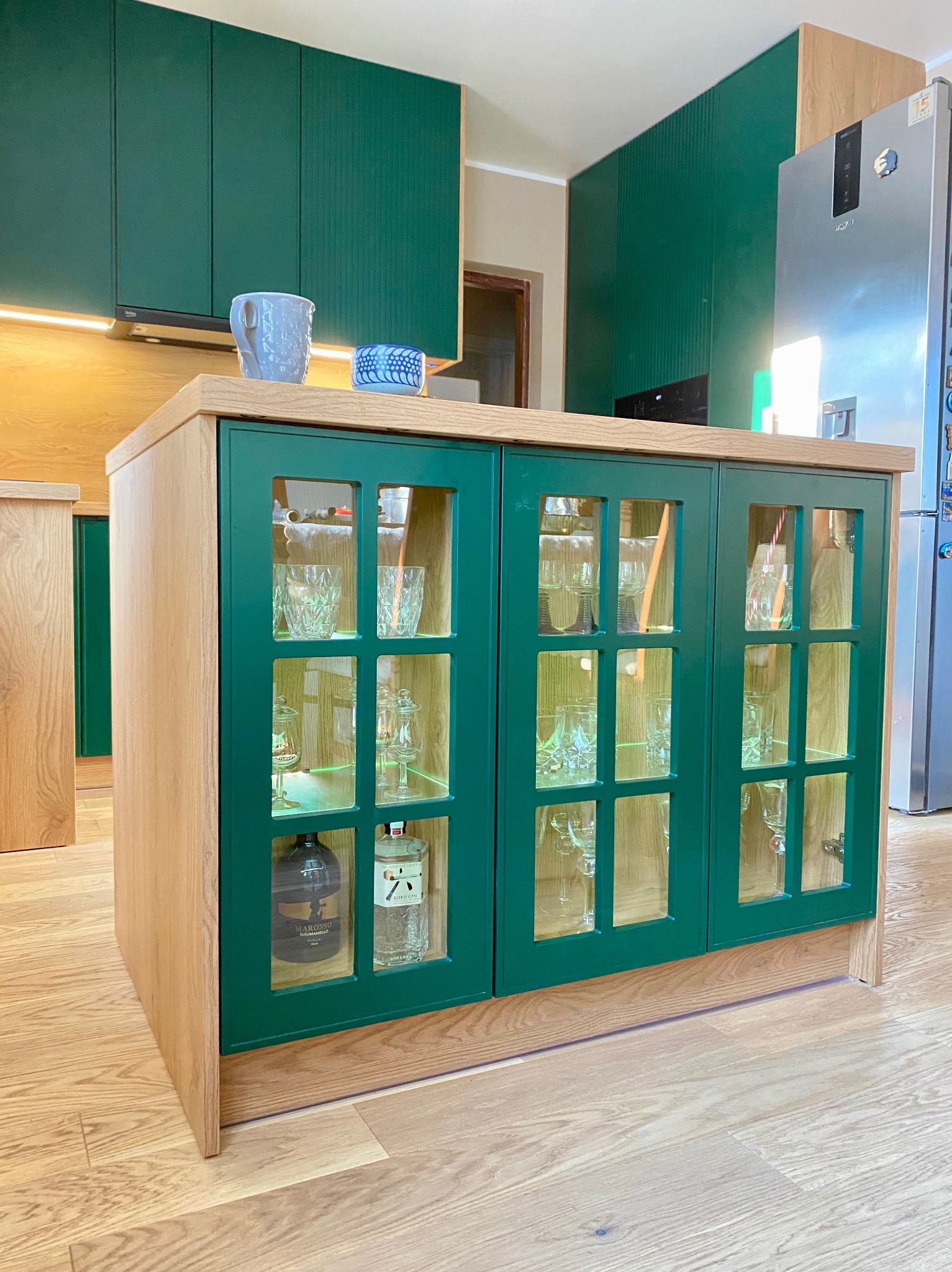 Zielona szafka z przeszklonymi drzwiczkami, podświetlona od wewnątrz, z ekspozycją szkła i alkoholi. Uzupełnienie aranżacji kuchni z zielonymi szafkami.