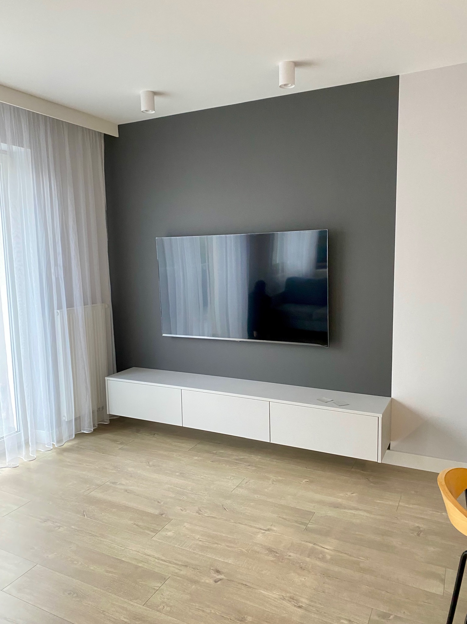 Minimalistyczny salon z wiszącą szafką RTV w bieli, pod telewizorem na szarej ścianie. Jasna podłoga i zasłony w oknie. Nowoczesne oświetlenie sufitowe.