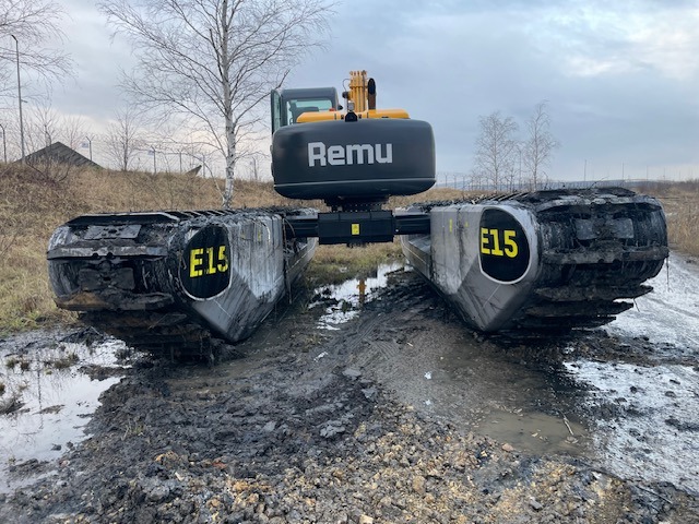 Amfibijna koparka gąsienicowa Remu E15 z tyłu, stojąca na błotnistym terenie. Widoczne gąsienice i fragment kabiny operatora. Pochmurne niebo w tle.