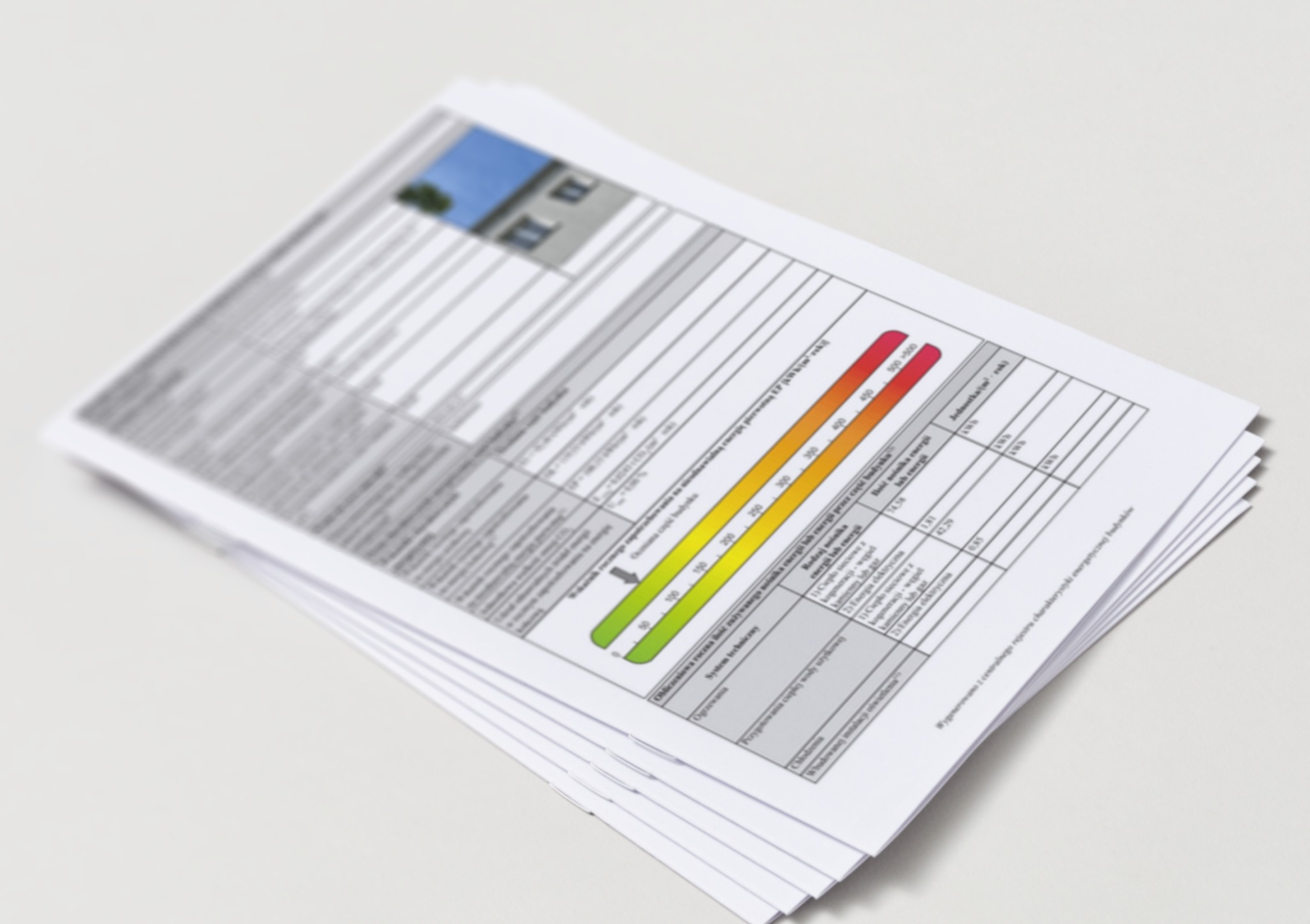 Stos dokumentów ze świadectwem energetycznym budynku, z widoczną skalą zużycia energii i danymi technicznymi. Dokumenty ułożone pod kątem na jasnym tle.