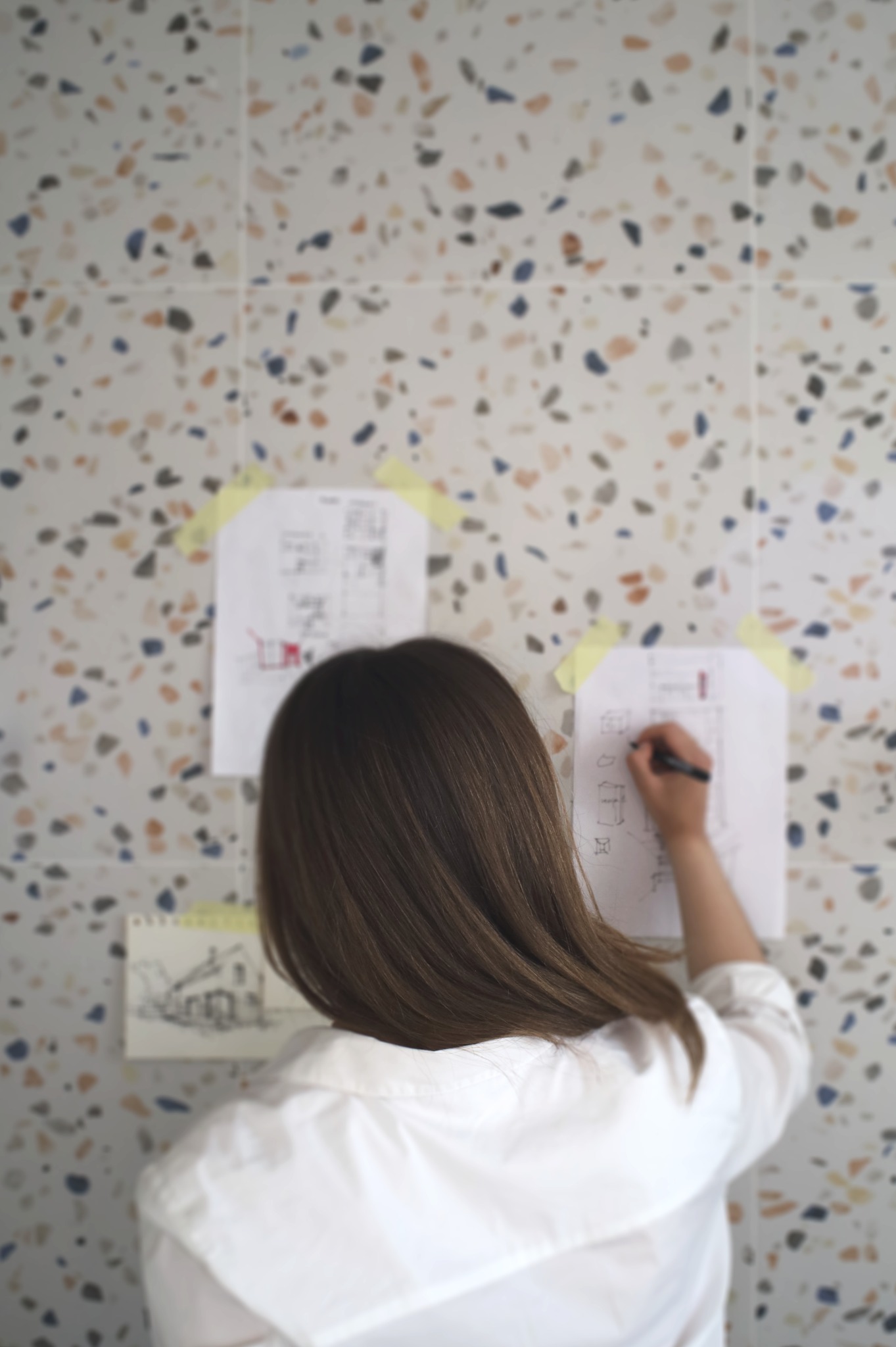 Kobieta w białej koszuli rysuje schemat na kartce przyklejonej do ściany w plamki. Widok z tyłu, skupienie na rysunku i ścianie.