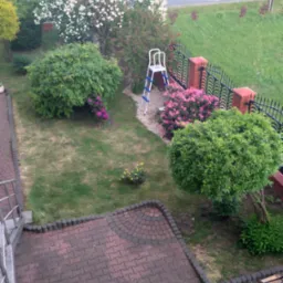 Ogród z perspektywy balkonu: kwitnące drzewa, krzewy różowe, trawnik, metalowe ogrodzenie z ceglanymi słupkami i drabina oparta o ogrodzenie.