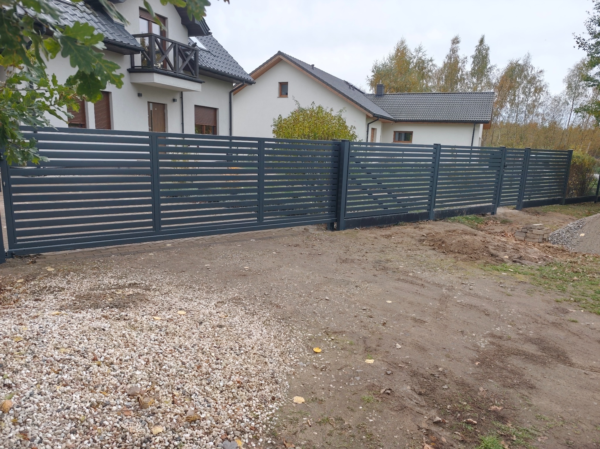 Nowoczesne, poziome ogrodzenie metalowe w kolorze grafitowym, z przesuwną bramą wjazdową. Tło stanowią dwa białe domy jednorodzinne i jesienne drzewa.