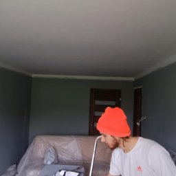 UNEXPERTED SPÓŁKA Z OGRANICZONĄ ODPOWIEDZIALNOŚCIĄ - Malowanie ścian na szaro w pokoju z listwami sufitowymi. Sofa zabezpieczona folią. Widoczny fragment osoby w pomarańczowej czapce podczas pracy.