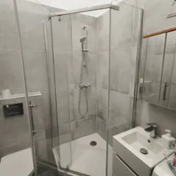 Szklana kabina prysznicowa z szarą armaturą i płytkami w nowoczesnej łazience. Widoczna umywalka z szafką, lustro i akcesoria łazienkowe.