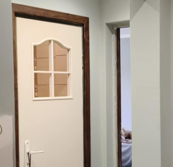 Zamknięte, białe drzwi z szybką w brązowej ramie, widoczne na tle jasnych ścian. Perspektywa z bliska, ukazująca detale wykończenia i kontrast kolorystyczny.