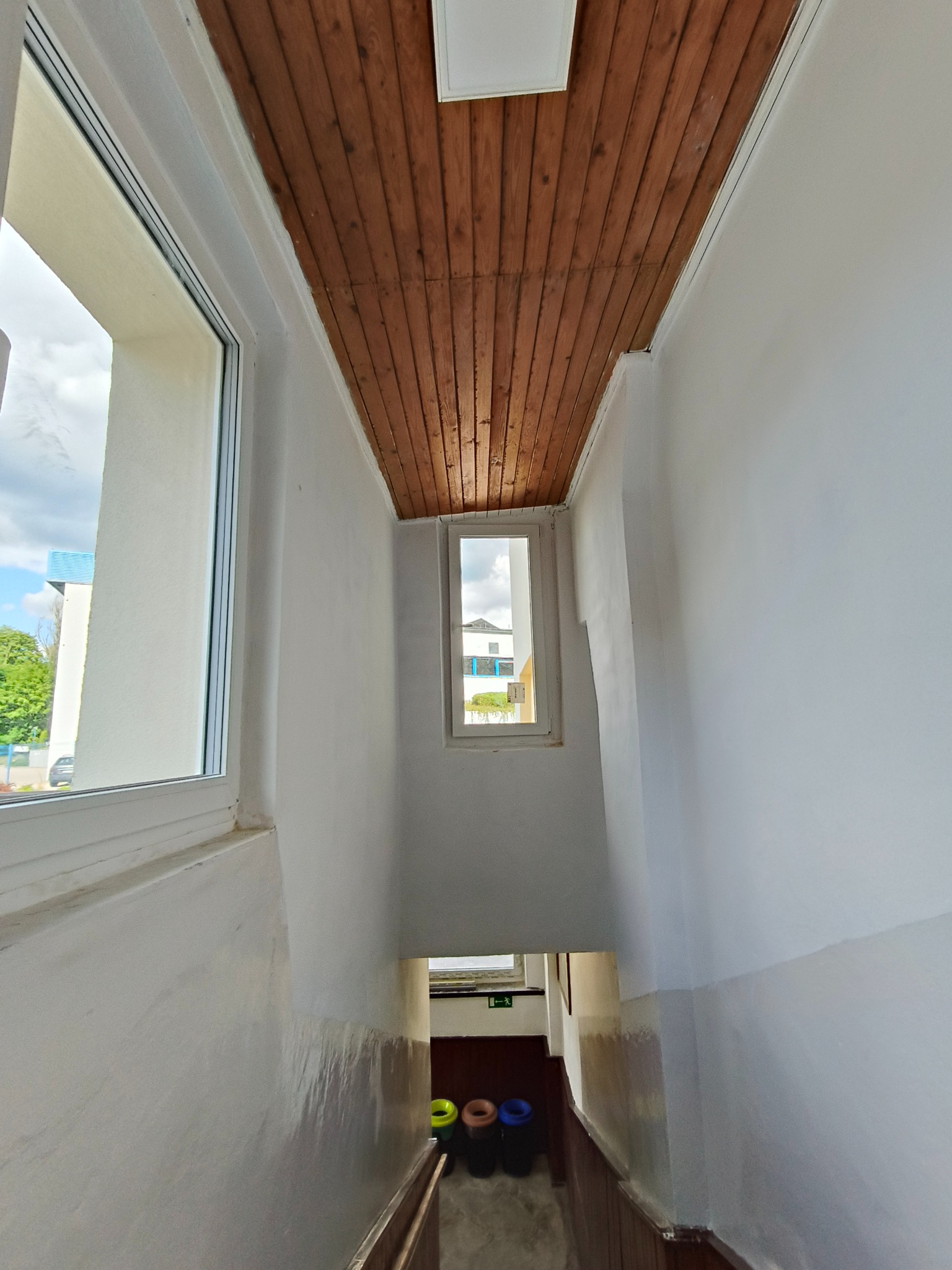 Klatka schodowa z drewnianym sufitem i oknami w budynku. Widoczne ślady malowania na ścianach i poręcz schodów. U dołu kosze na śmieci.