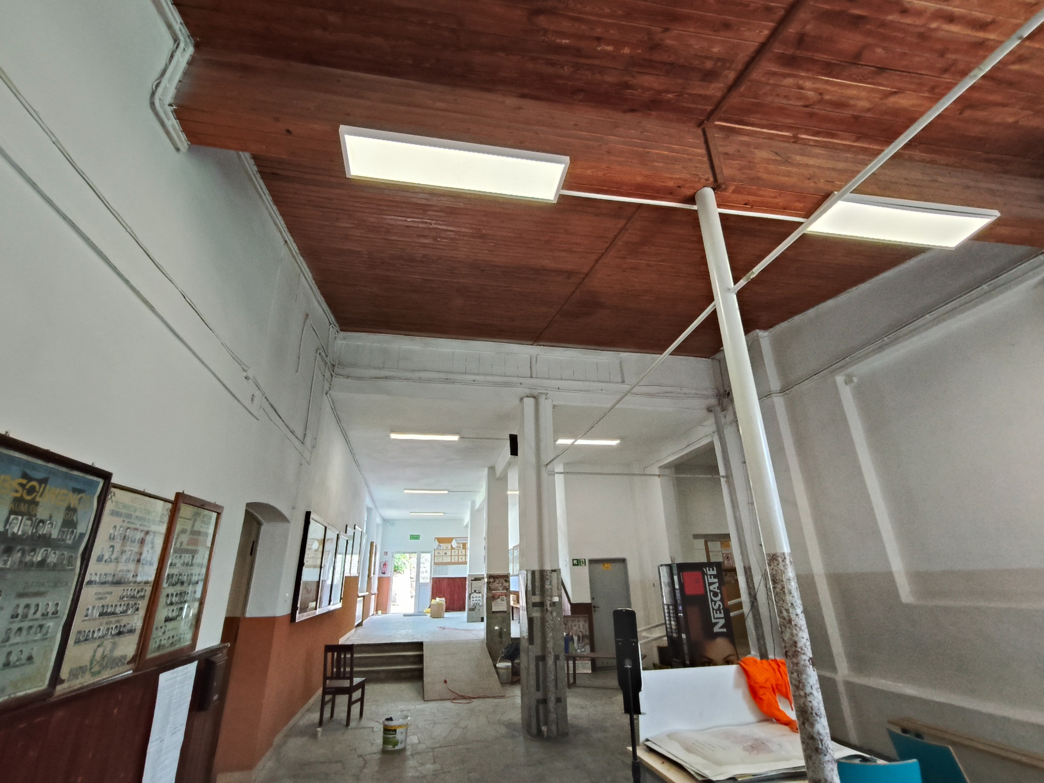Wnętrze korytarza po remoncie: białe ściany, drewniany sufit z lampami LED, tablice ze zdjęciami na ścianach. Widok w głąb korytarza, maszyna Nescafé.