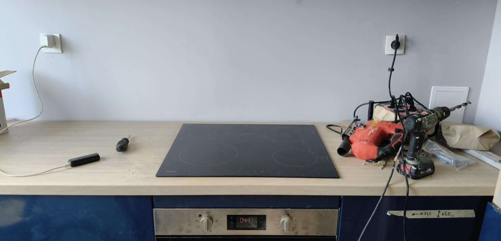 Montaż płyty indukcyjnej na blacie kuchennym w trakcie prac. Widoczne narzędzia: wiertarka, szlifierka, śrubokręt. Niebieskie szafki kuchenne i szara ściana w tle.