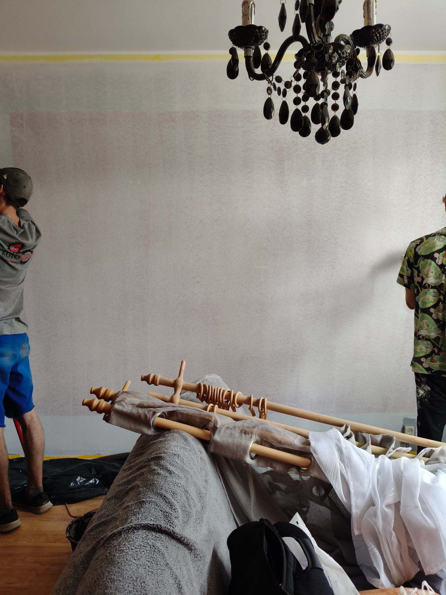 Dwóch mężczyzn maluje ścianę w pokoju z żyrandolem. Widoczne wałki, drabina i zabezpieczona taśmą górna krawędź ściany.