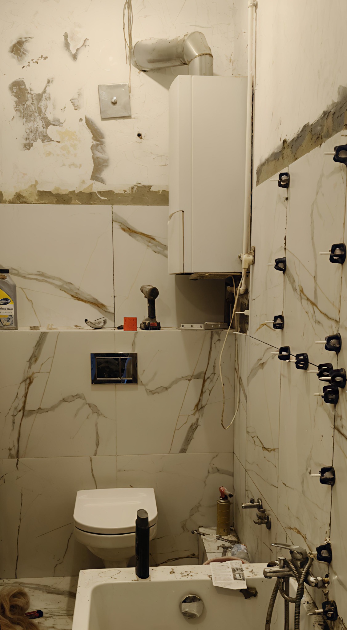Remont łazienki w trakcie: białe płytki z marmurkowym wzorem, wisząca toaleta, bojler. Widoczne narzędzia i system poziomowania płytek na ścianie.