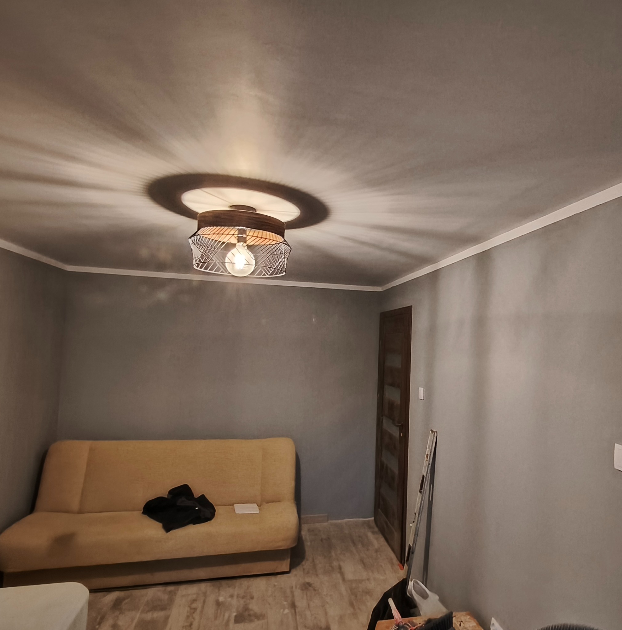 Szpachlowane ściany w szarym odcieniu z dekoracyjną lampą sufitową, beżowa sofa i widoczne narzędzia malarskie. Wnętrze po remoncie.