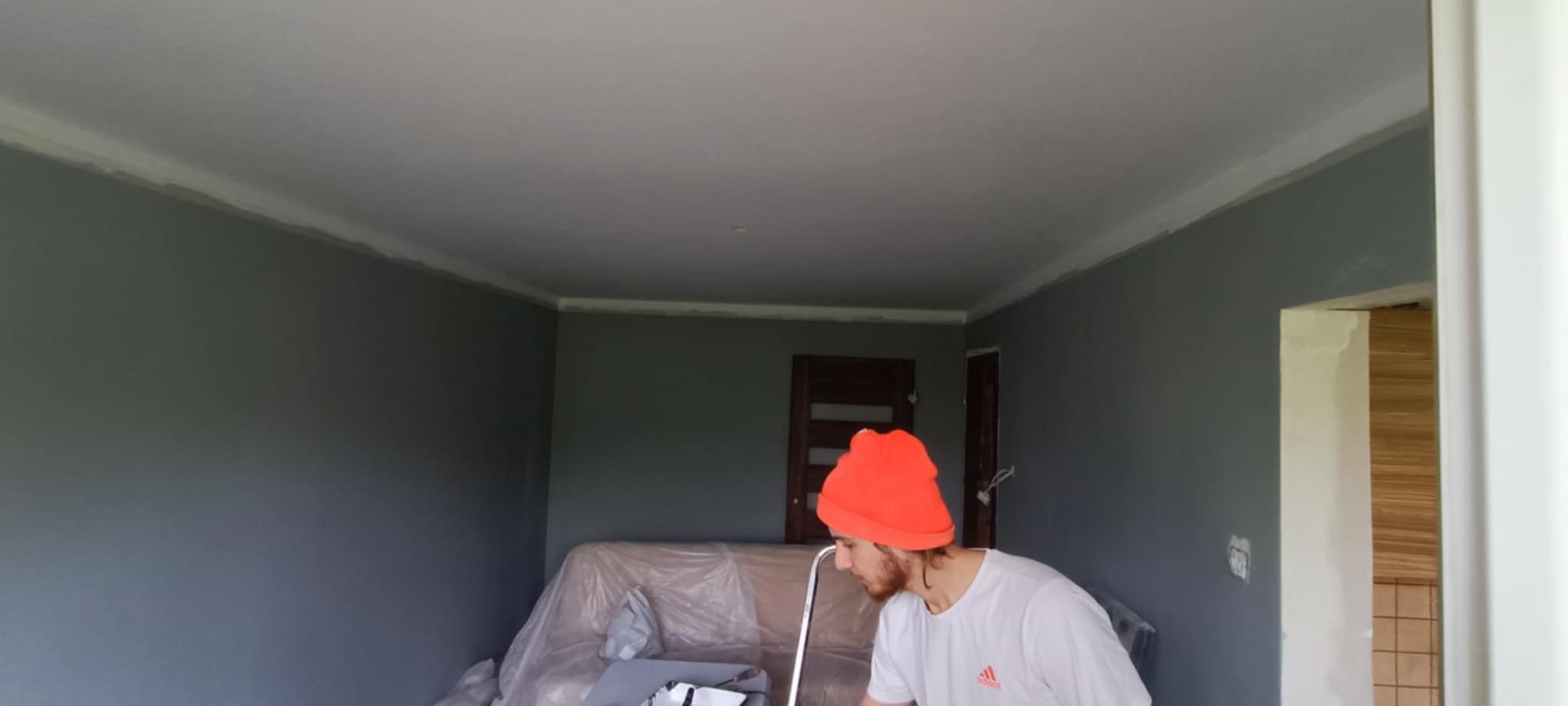 Malowanie ścian na szaro w pokoju z listwami sufitowymi. Sofa zabezpieczona folią. Widoczny fragment osoby w pomarańczowej czapce podczas pracy.