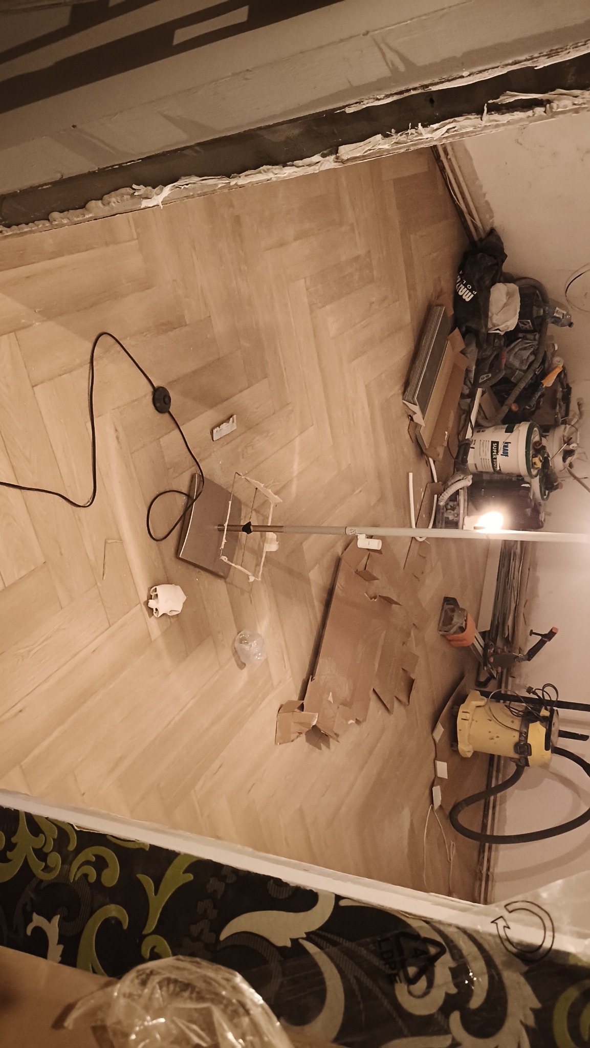 Parkiet ułożony w jodełkę, przygotowany do wykończenia. Na podłodze narzędzia, kartony i odkurzacz. Widoczne niedokończone wykończenia ścian.