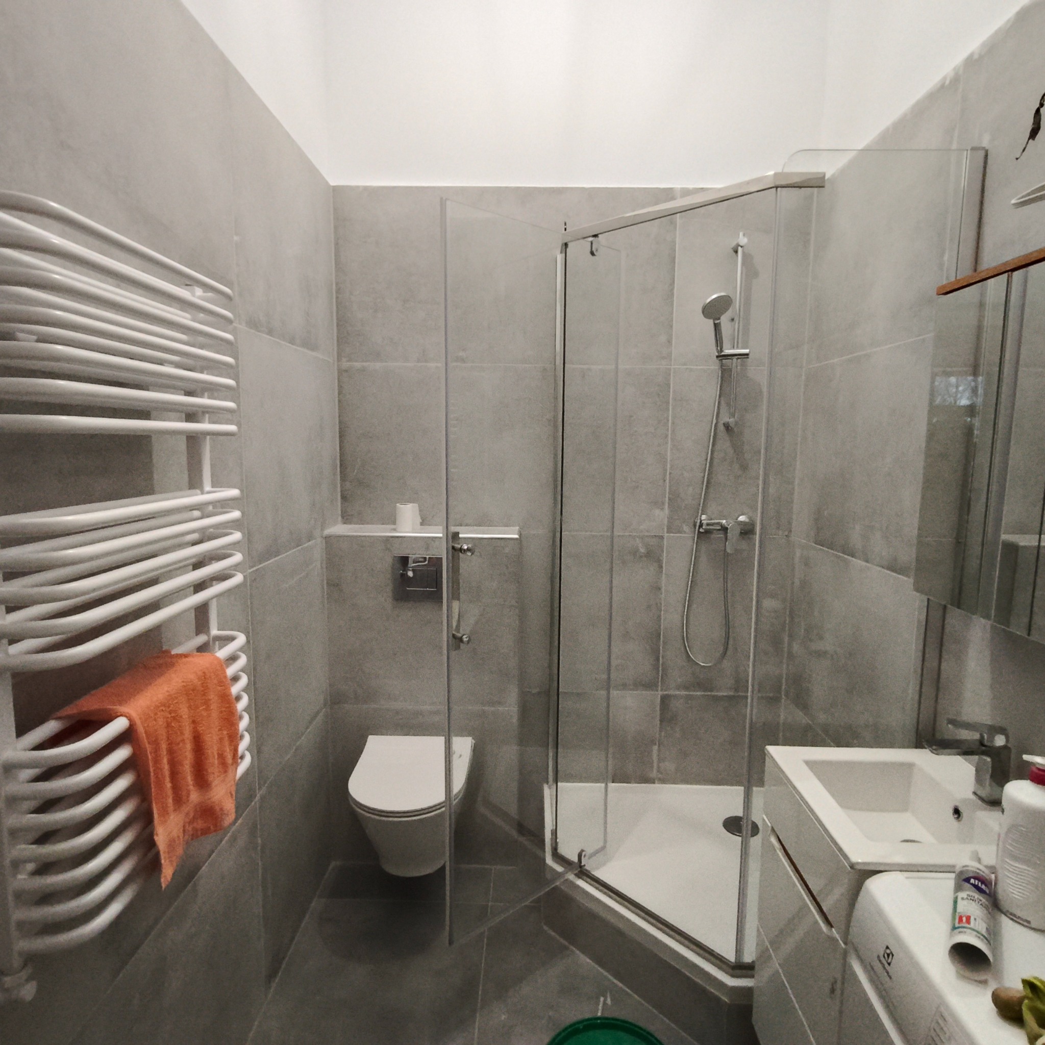 Wykończona łazienka w szarościach: kabina prysznicowa z brodzikiem, toaleta podtynkowa, umywalka z szafką i grzejnik drabinkowy z pomarańczowym ręcznikiem.