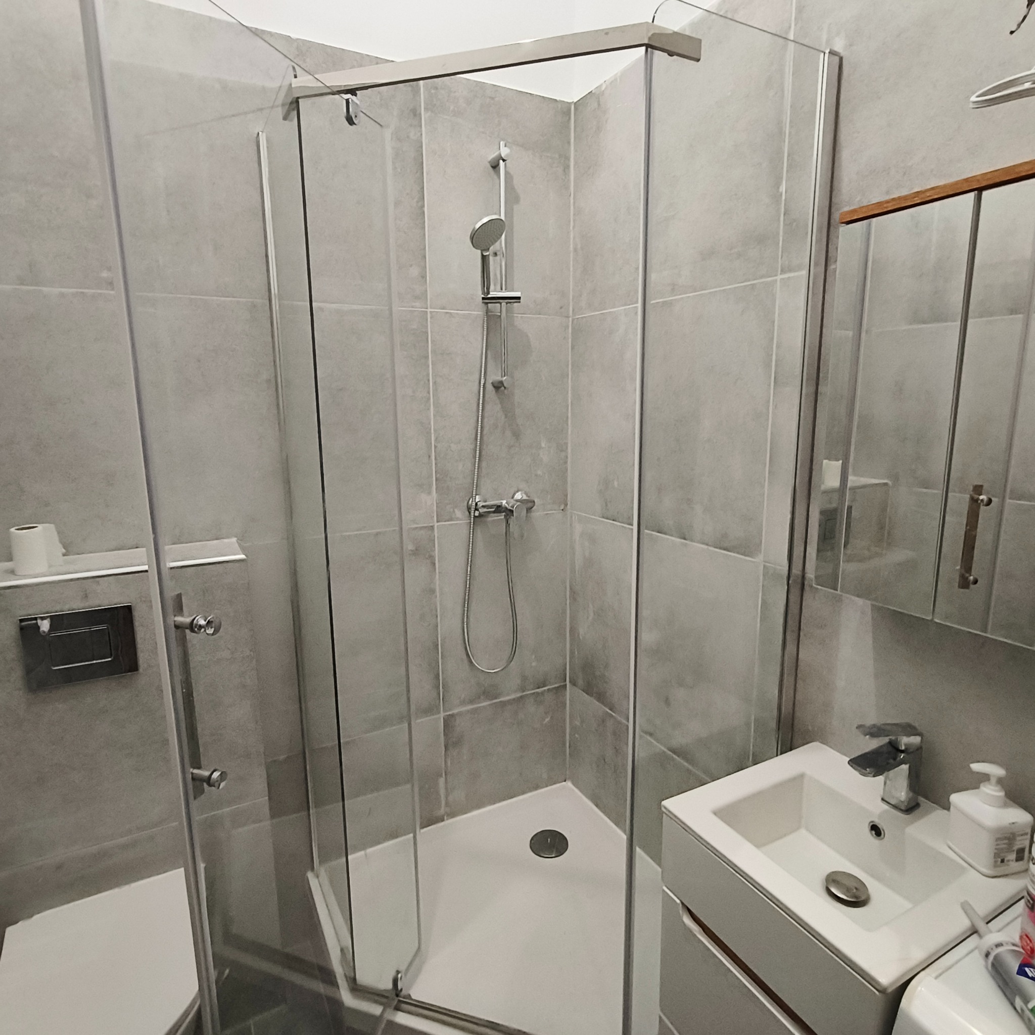 Szklana kabina prysznicowa z szarą armaturą i płytkami w nowoczesnej łazience. Widoczna umywalka z szafką, lustro i akcesoria łazienkowe.