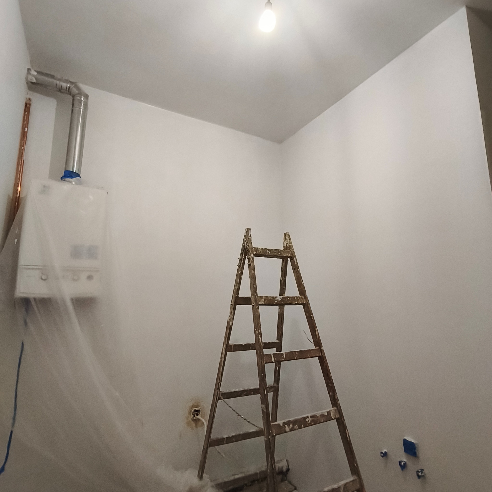 Białe, świeżo pomalowane wnętrze z drewnianą drabiną, zabezpieczonym piecem gazowym i widocznymi puszkami instalacji elektrycznej. Praca w toku.