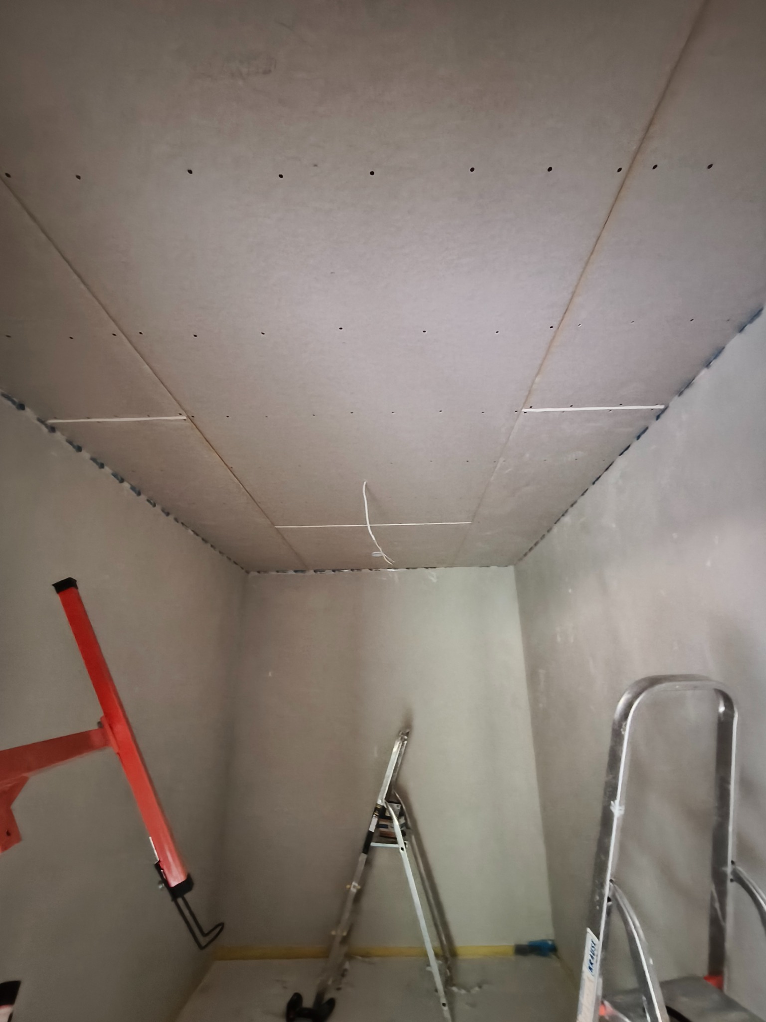 Wnętrze pokoju w trakcie montażu płyt karton-gips na suficie. Widoczne drabiny i fragment instalacji elektrycznej. Ściany przygotowane pod szpachlowanie.