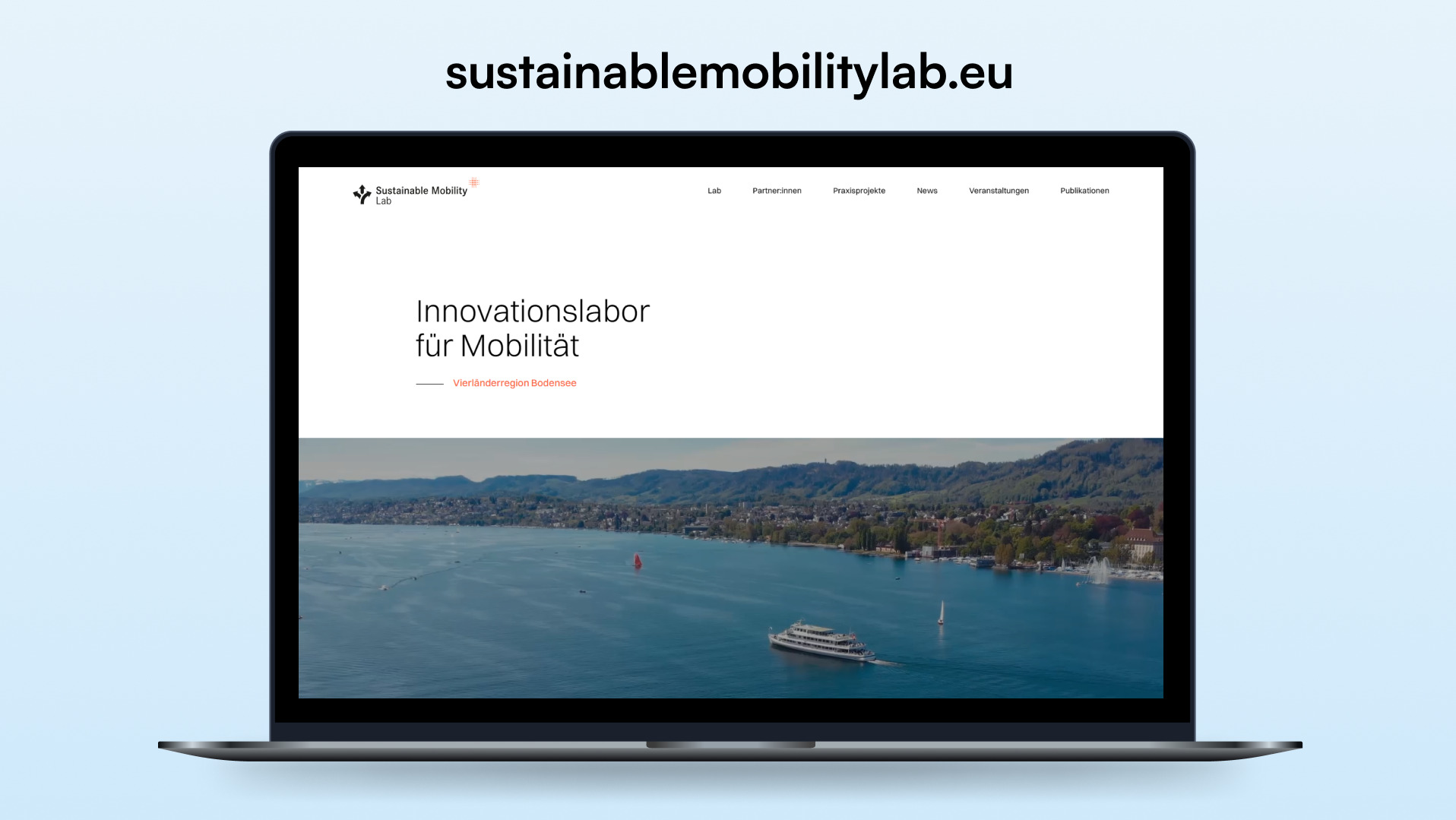 Ekran laptopa prezentuje stronę internetową sustainablemobilitylab.eu, z widokiem na jezioro i góry w tle. Minimalistyczny design, białe tło i niemiecki tekst.