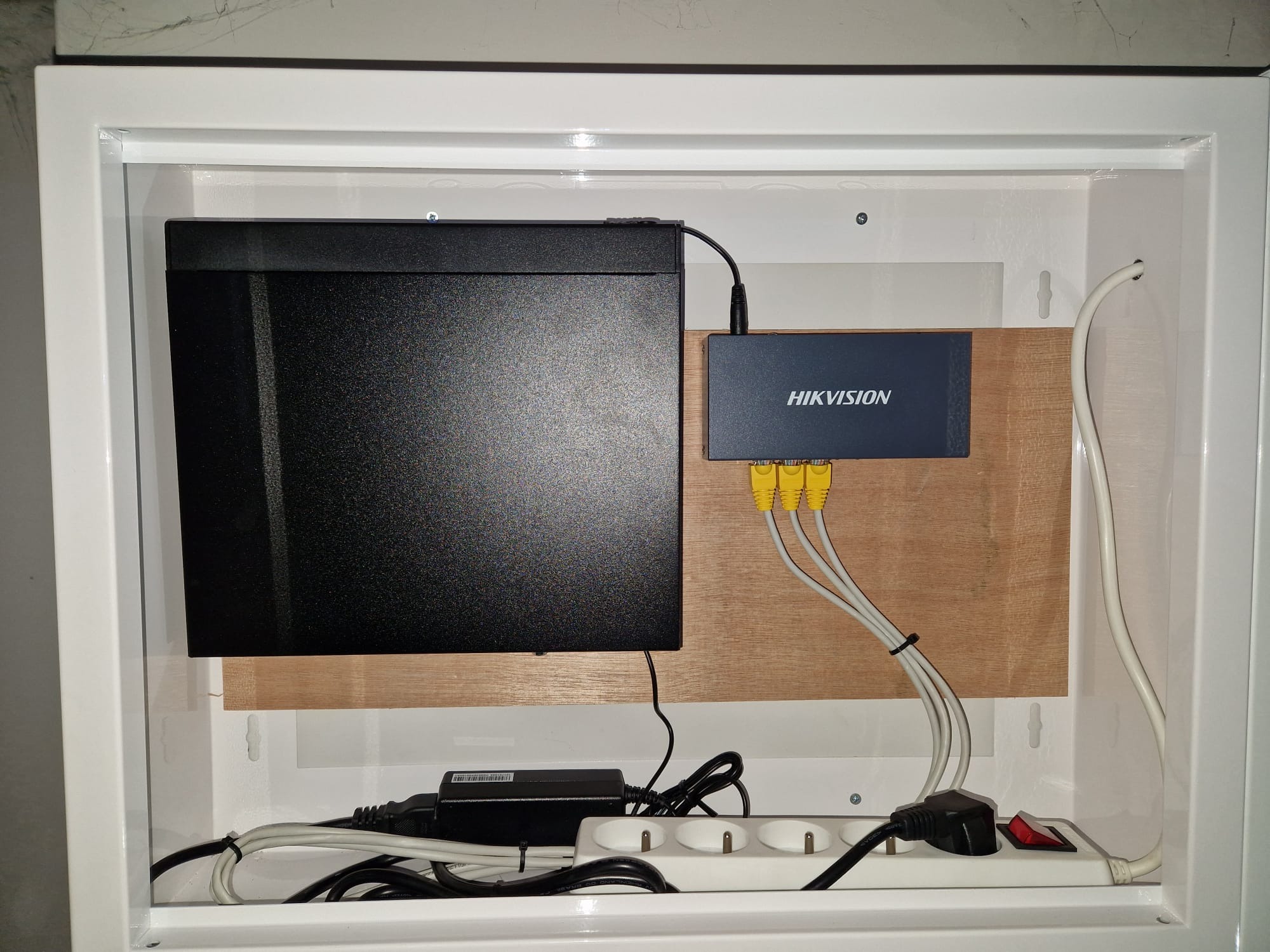 Zamknięta szafka z zamontowanym rejestratorem i routerem marki Hikvision, z podłączonymi kablami Ethernet i zasilaniem. Widoczna listwa zasilająca.