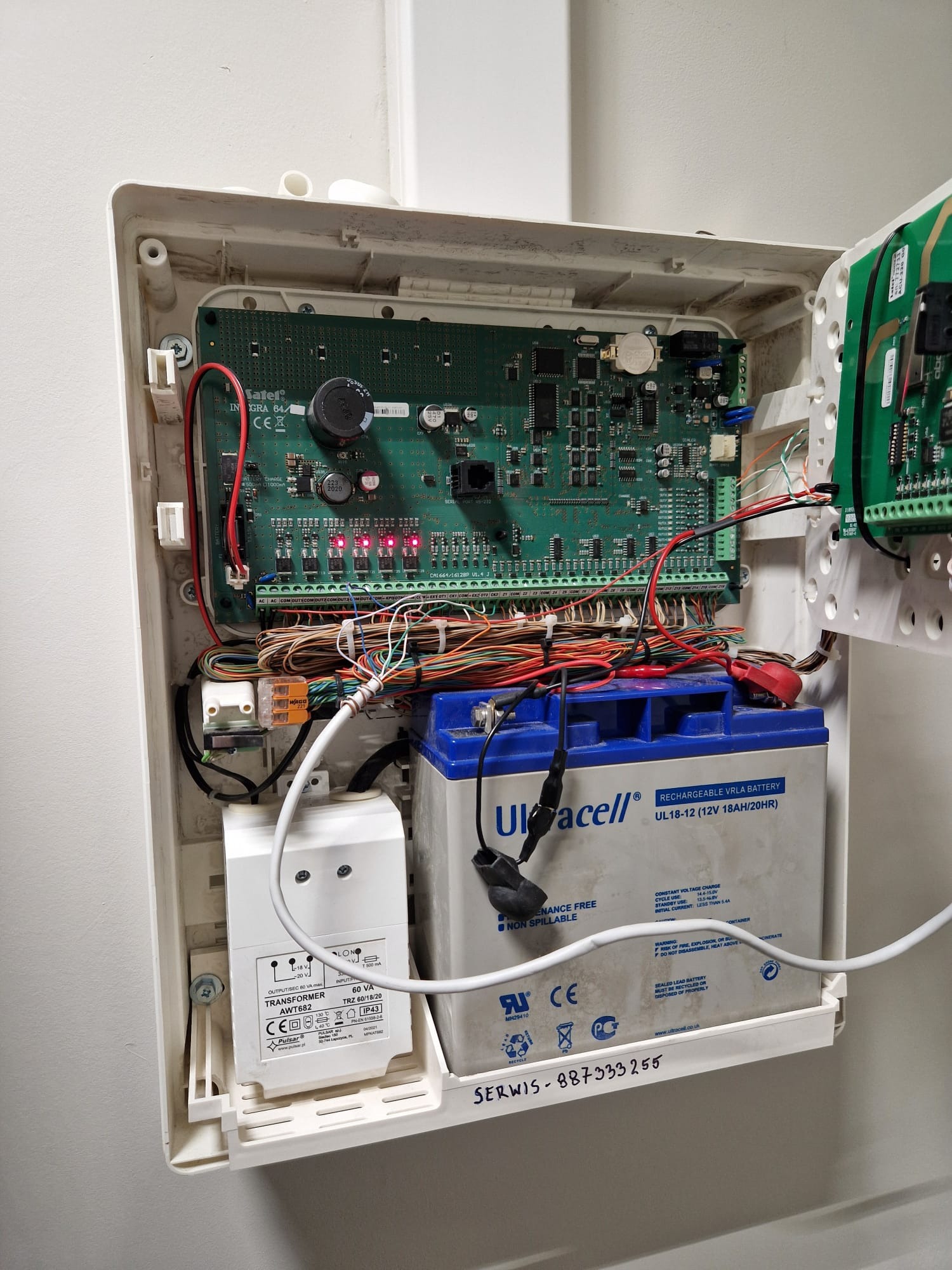 Otwarta obudowa centrali alarmowej z widoczną elektroniką, akumulatorem i transformatorem. Zapisany numer serwisu na obudowie.