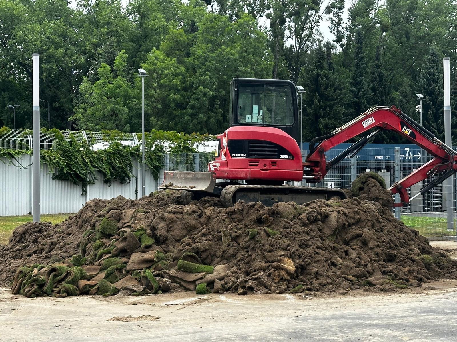 Czerwona koparka Zeppelin 305D na stercie ziemi z fragmentami darni, na tle ogrodzenia i zieleni. Prace ziemne w otoczeniu miejskim.