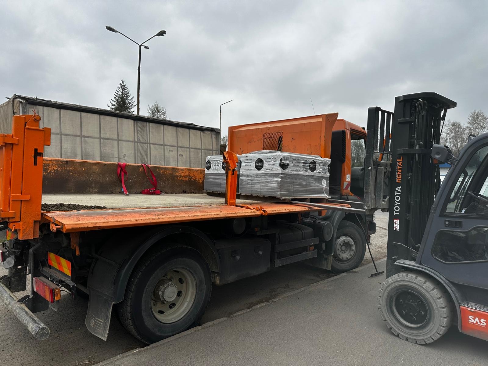 Pomarańczowa ciężarówka z kostką brukową i wózkiem widłowym w Sosnowcu. Widoczne palety z kostką Certus, gotowe do rozładunku. Transport materiałów budowlanych.