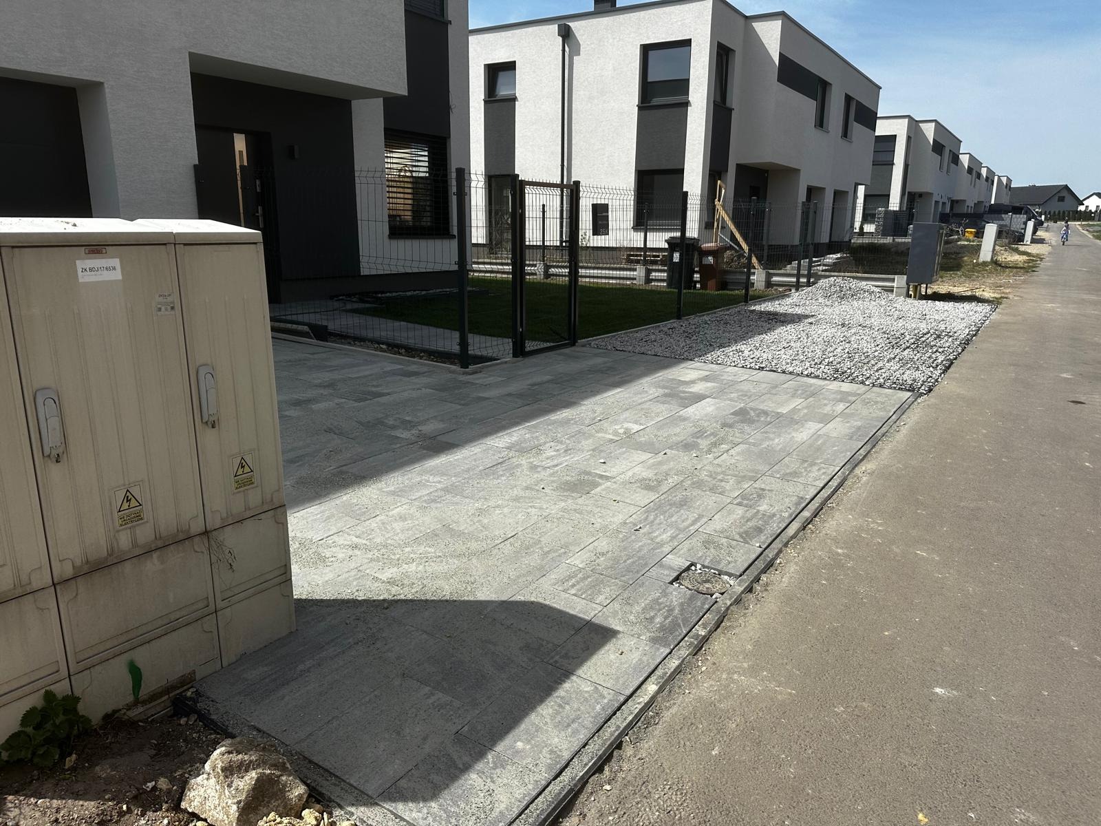 Szara kostka brukowa na osiedlu domów szeregowych, widok z poziomu gruntu. W tle siatka ogrodzeniowa i biały budynek. Widoczna studzienka rewizyjna.