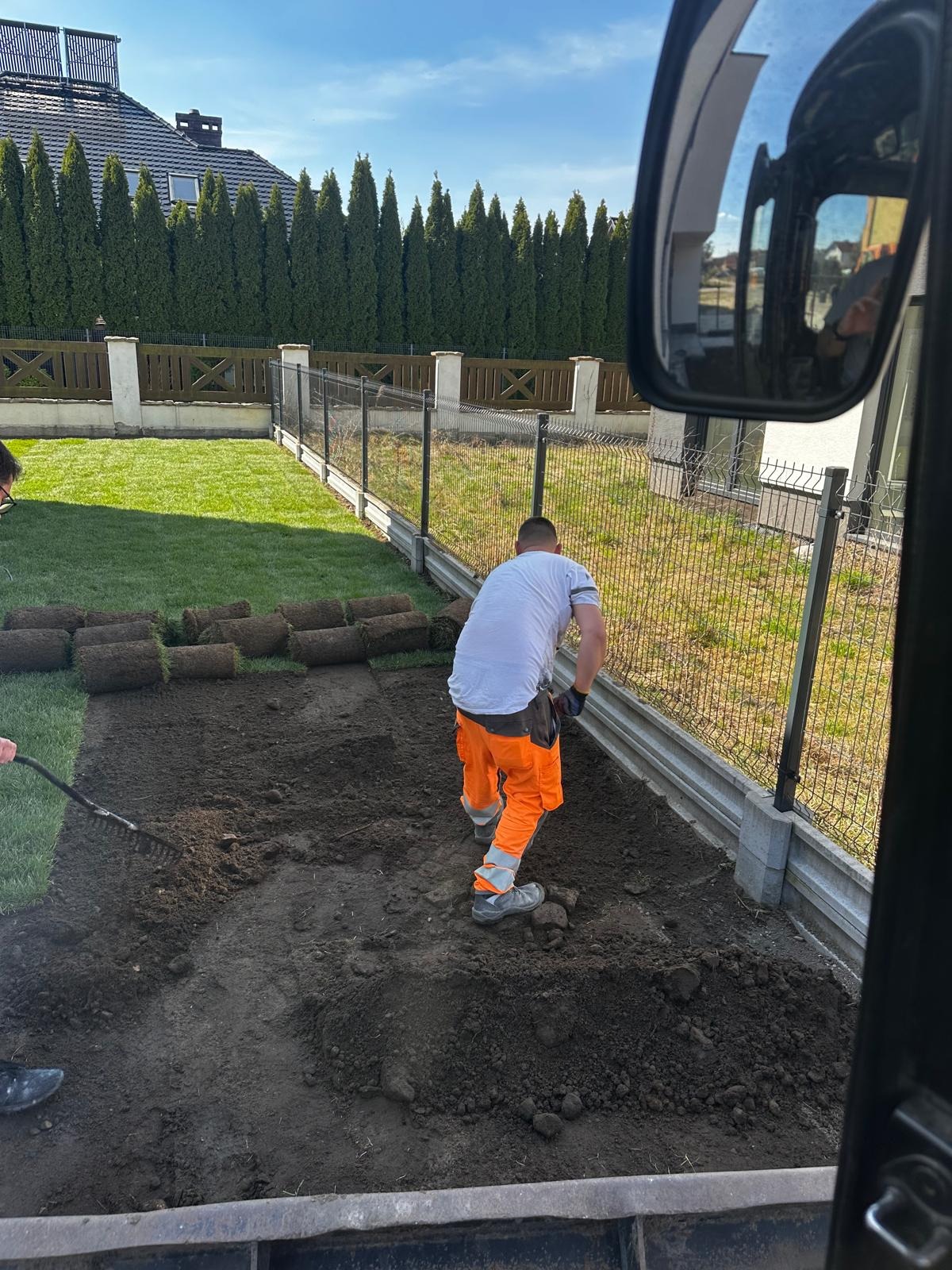Pracownik w pomarańczowych spodniach rozkłada darń na przygotowanym terenie przy ogrodzeniu. Widok z kabiny maszyny na trawnik i tuje.
