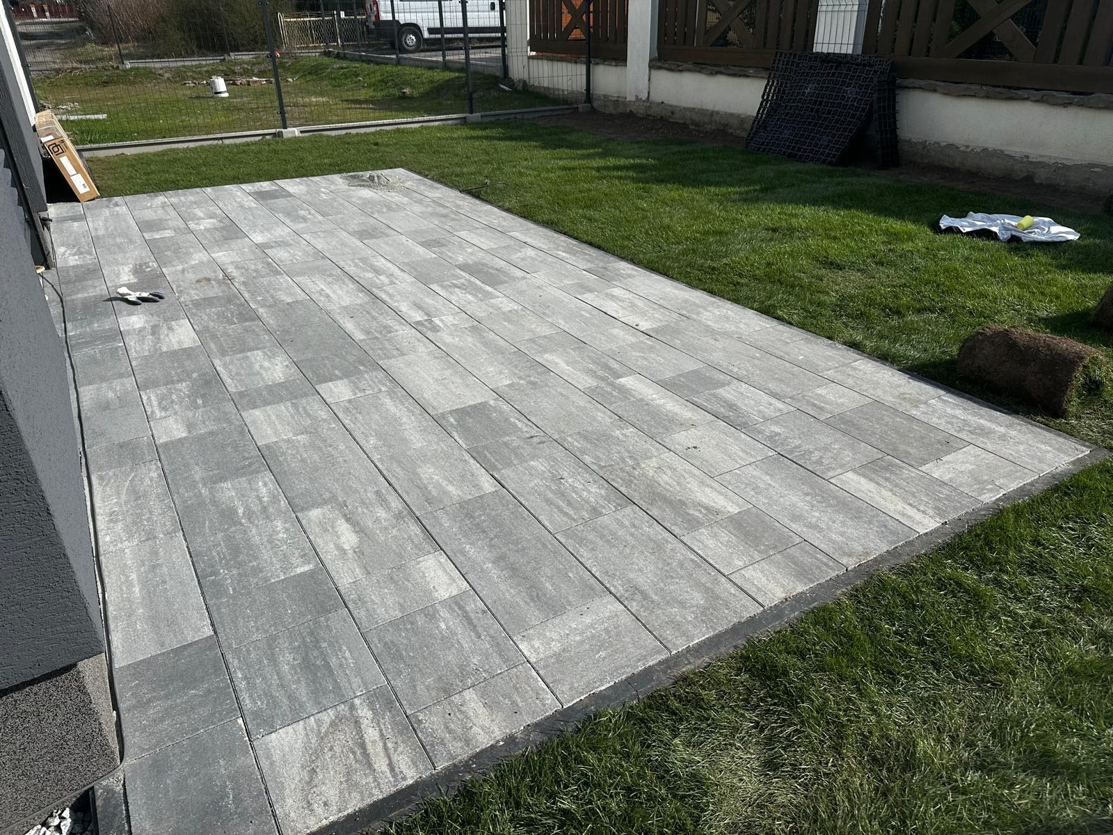 Ułożona, szara kostka brukowa na trawniku, przylegająca do ściany budynku. Widoczne rolki trawy i akcesoria, sugerujące świeżo zakończone prace.
