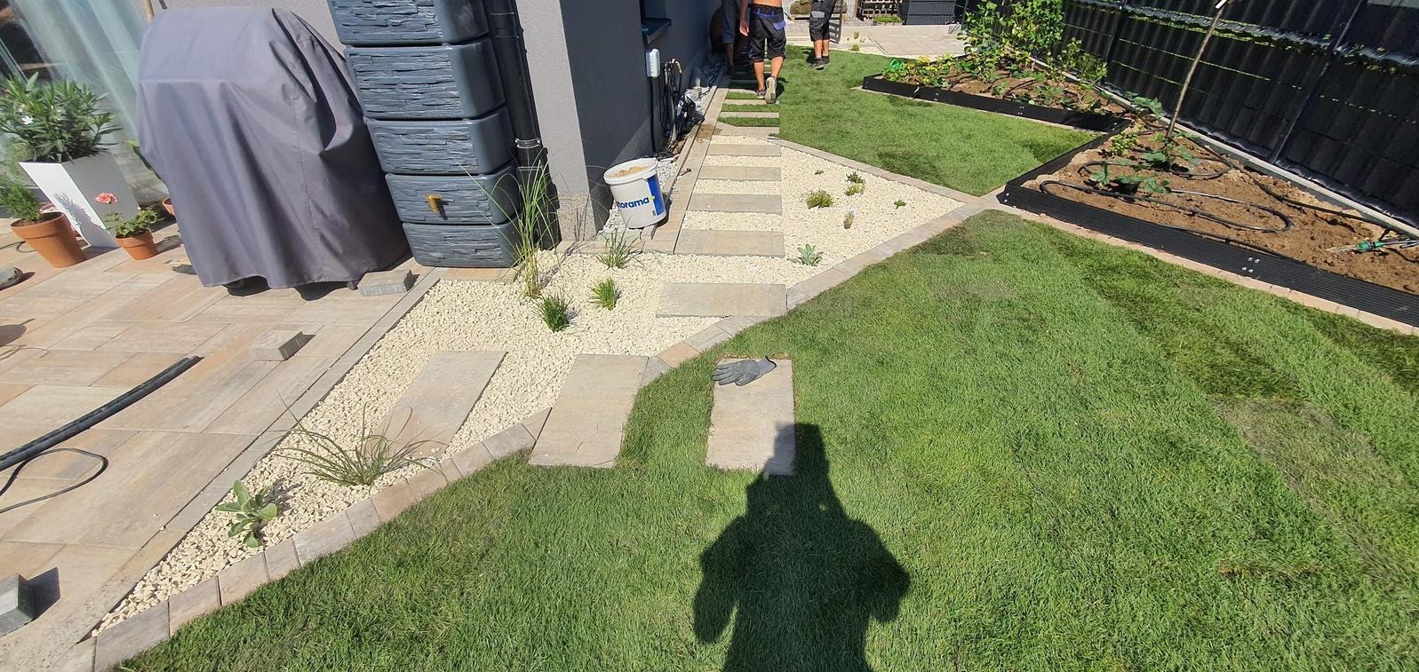 Ścieżka z płyt i białego żwiru w ogrodzie z trawnikiem i rabatami. Widoczny zbiornik na deszczówkę i cień fotografa. Ułożona kostka brukowa.