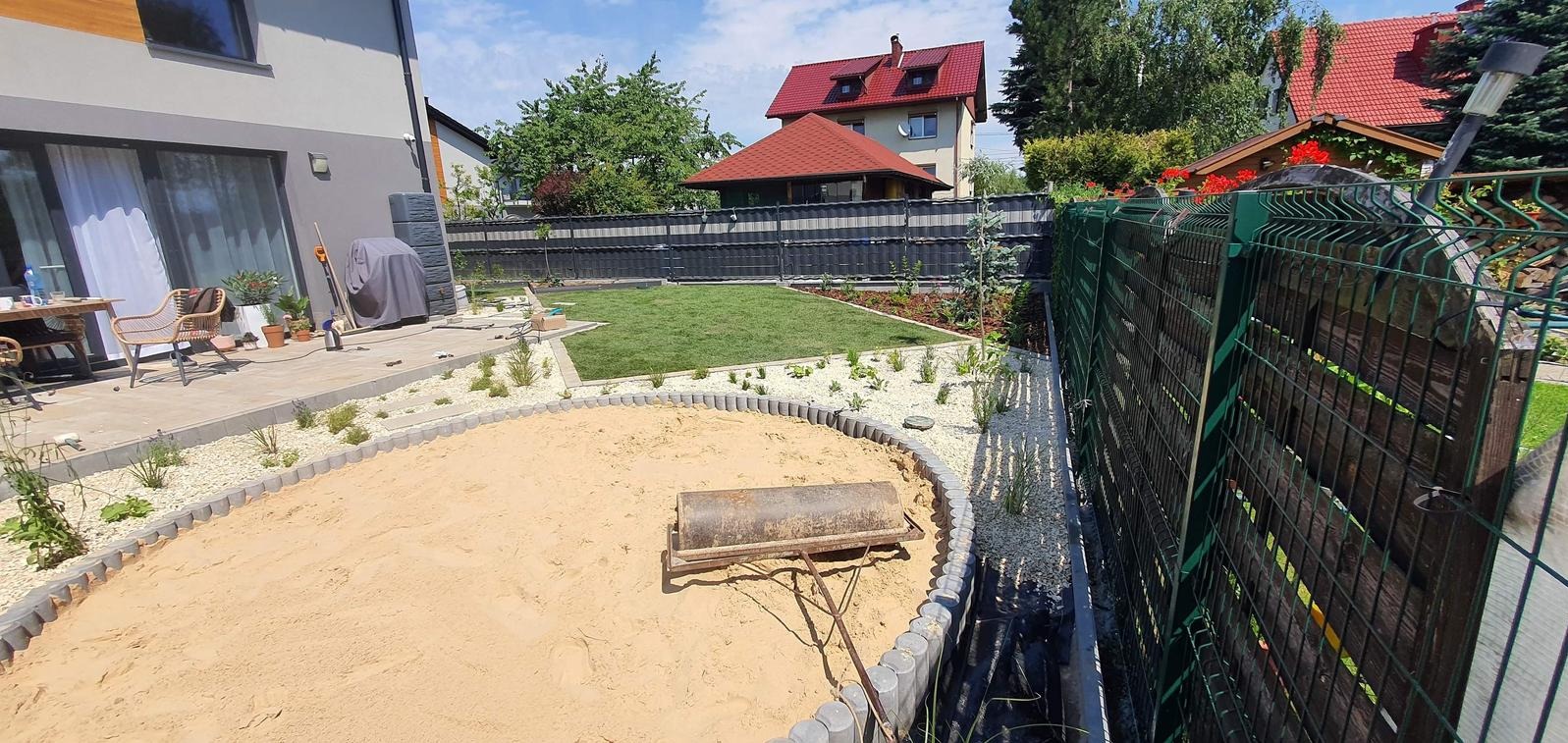 Ogród po częściowej renowacji: piaskownica z obrzeżem, trawnik, rabaty z kamykami i roślinami, nowe ogrodzenie panelowe. Walec ogrodowy na piasku.