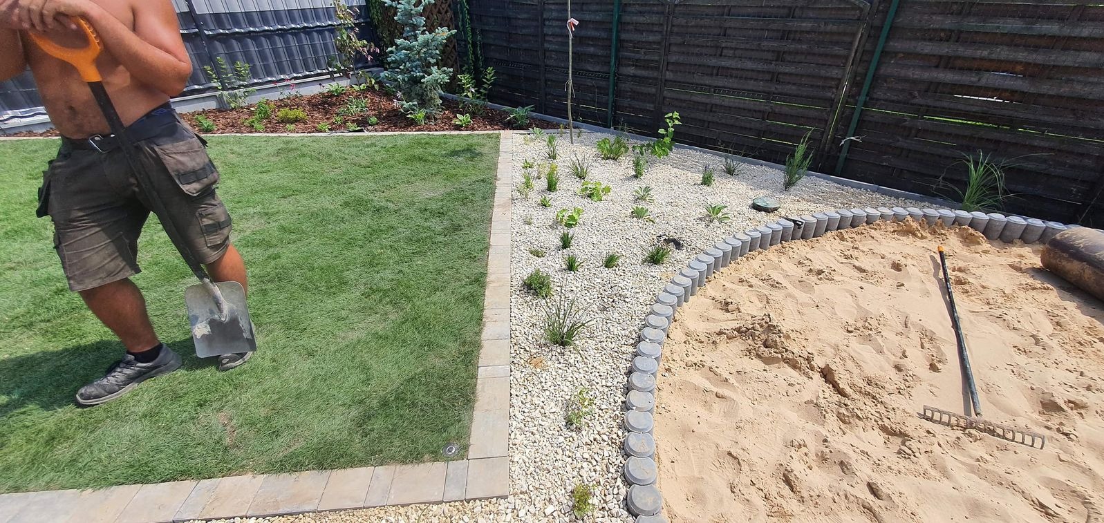 Ukończony ogród: trawnik, rabaty z białym żwirem i roślinami, obrzeże piaskownicy z kostki. Mężczyzna z łopatą na trawniku. Drewniany płot w tle.