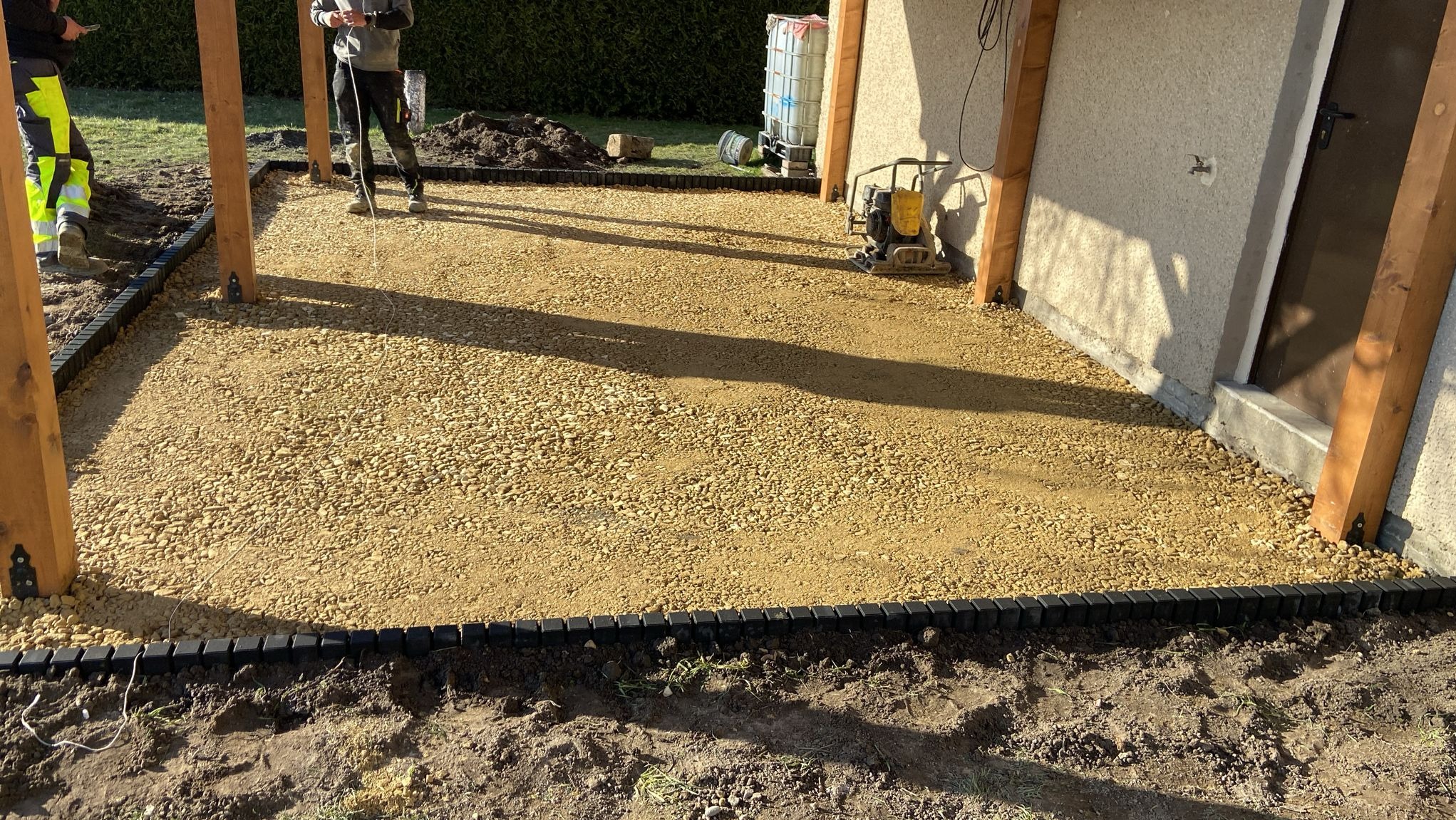 Przygotowany teren pod kostkę brukową z obrzeżami i ubijarką w tle. Widoczni pracownicy podczas niwelacji terenu podłoża w słoneczny dzień.