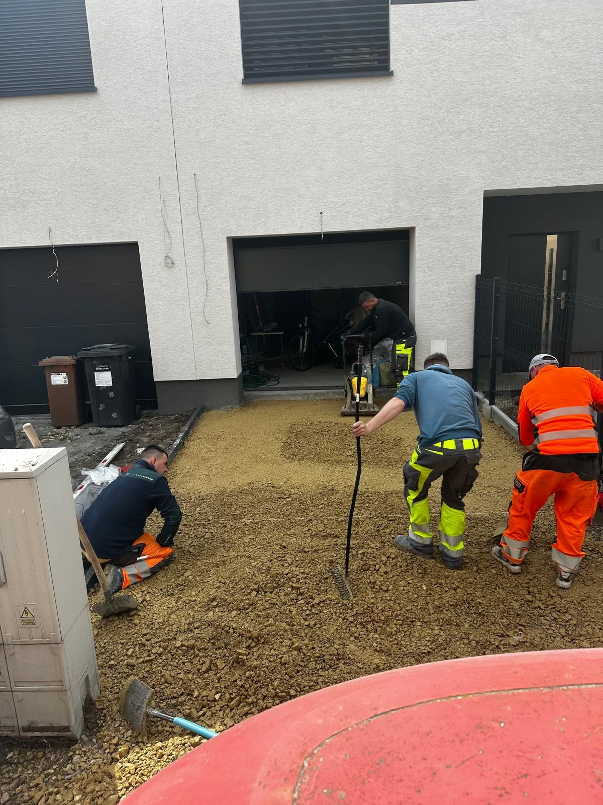 Ekipa w odblaskowych strojach wyrównuje podłoże pod kostkę brukową na posesji z garażem. W tle narzędzia i maszyna do ubijania.