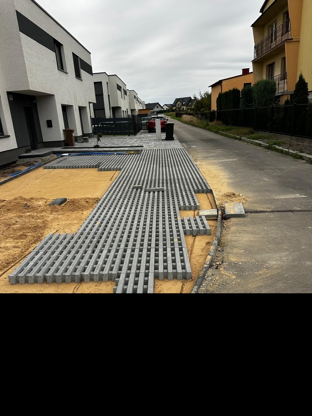 Fragment układania ażurowej kostki brukowej na piasku, widoczne budynki mieszkalne w tle. Częściowo ułożona nawierzchnia z kostki.
