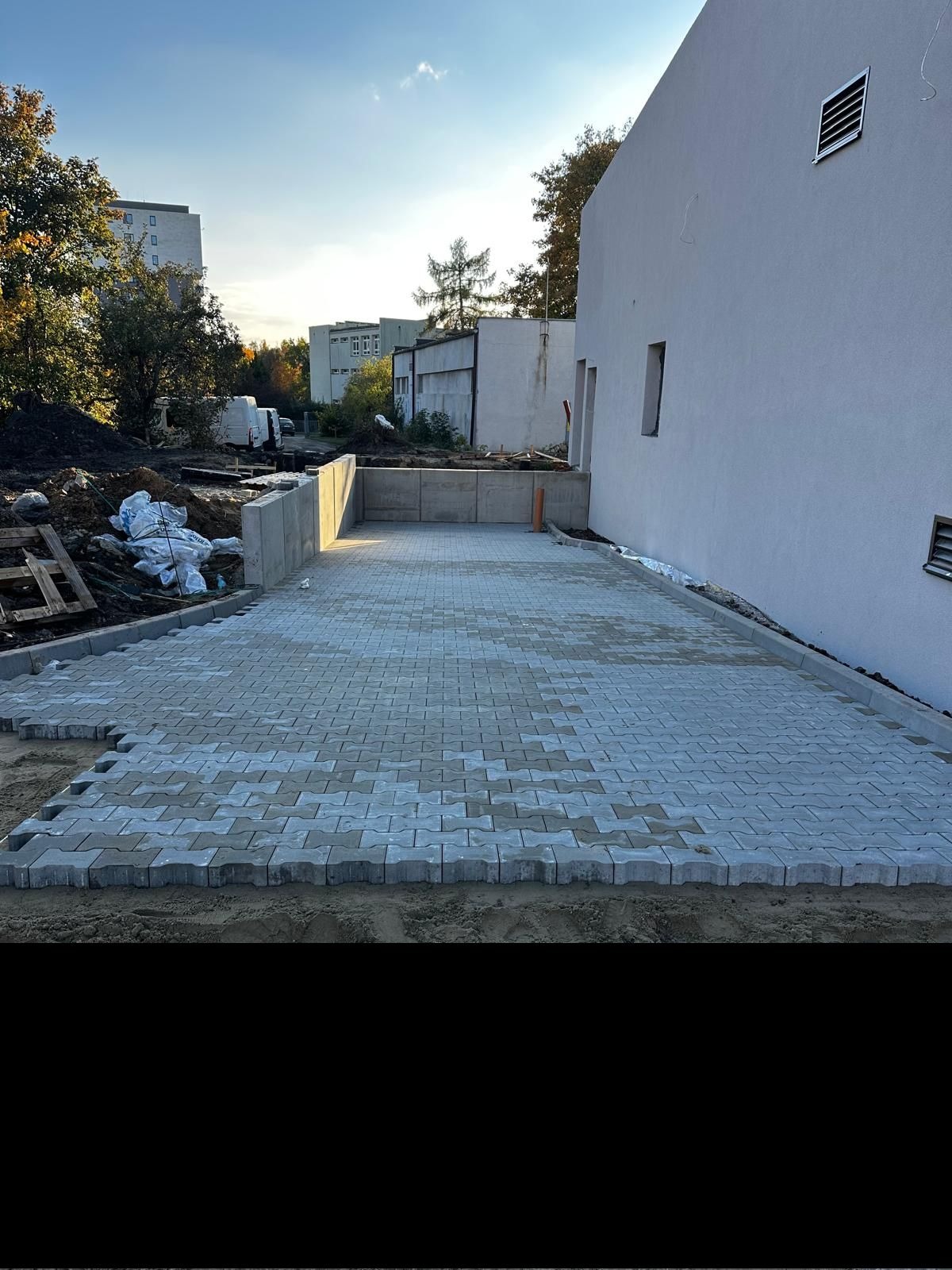 Fragment układania szarej kostki brukowej typu 'psia kość' na podsypce piaskowej, widoczne krawężniki i przygotowany teren pod dalsze prace.