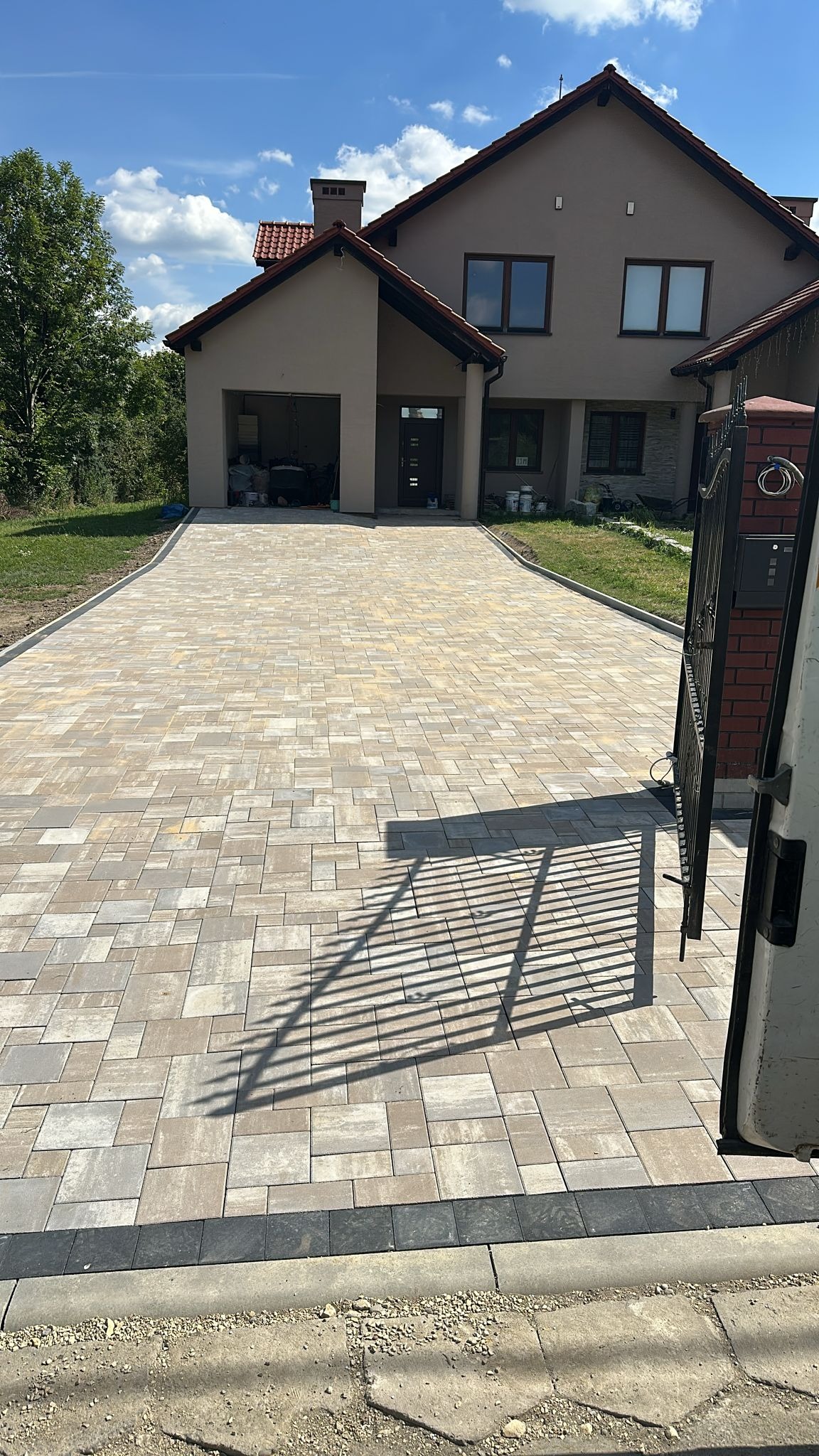 Ułożona kostka brukowa w odcieniach beżu i szarości tworzy wzór na podjeździe przed domem z garażem. Cień bramy dodaje głębi. Widoczne obrzeża i trawnik.